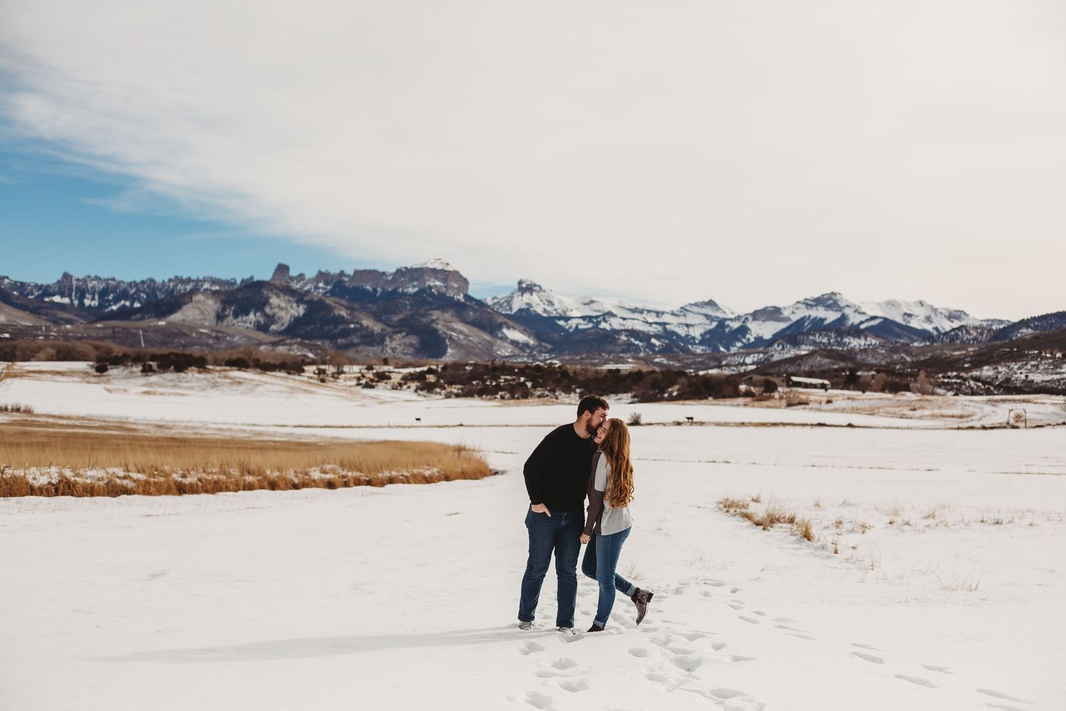 A couple walks together on snowy terrain with snow-capped mountains in the background on a sunny winter day.