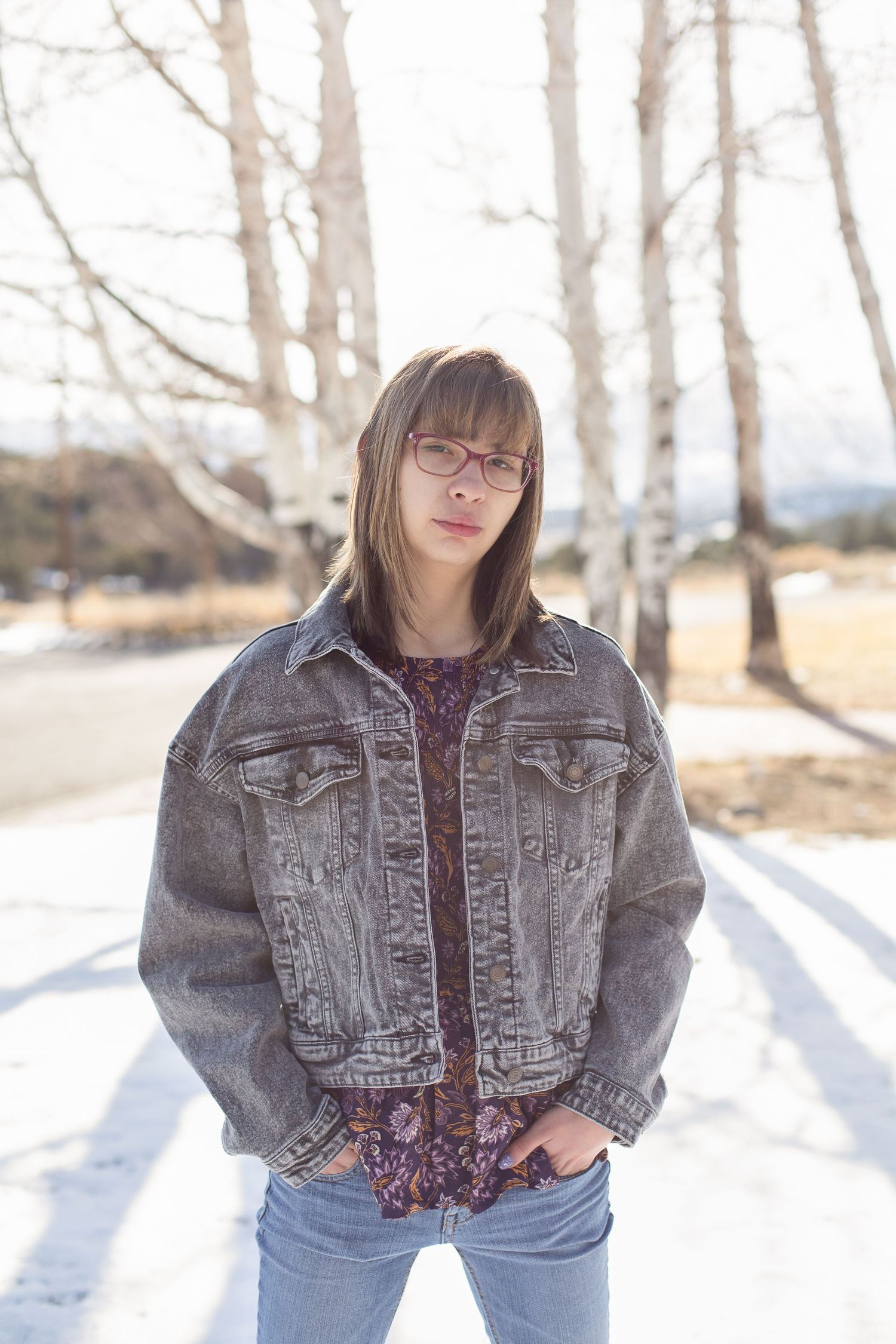 Person in denim jacket standing on snowy road with birch trees in background.