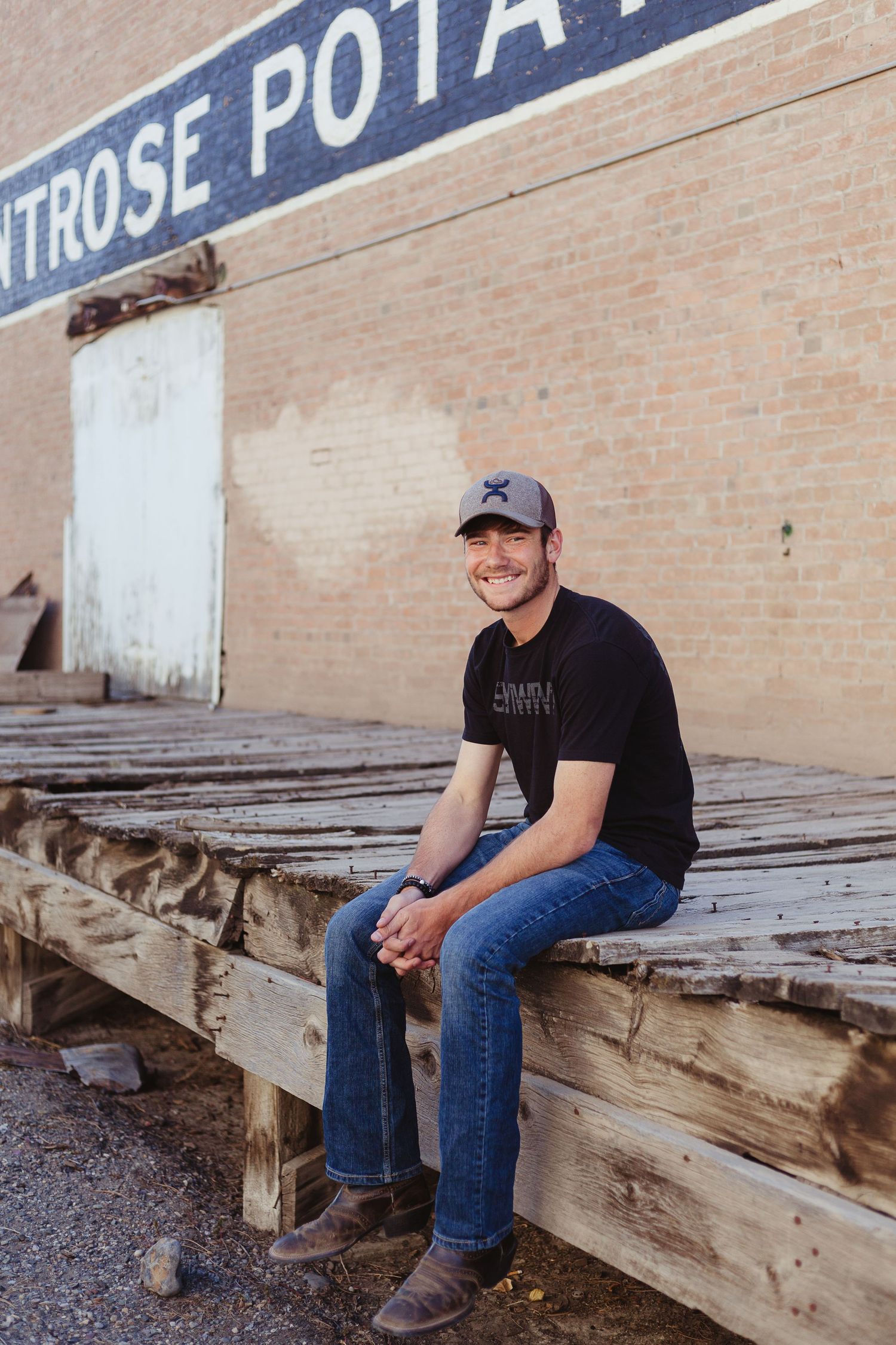 Person in black t-shirt and jeans sitting on rustic wooden seating against brick wall.