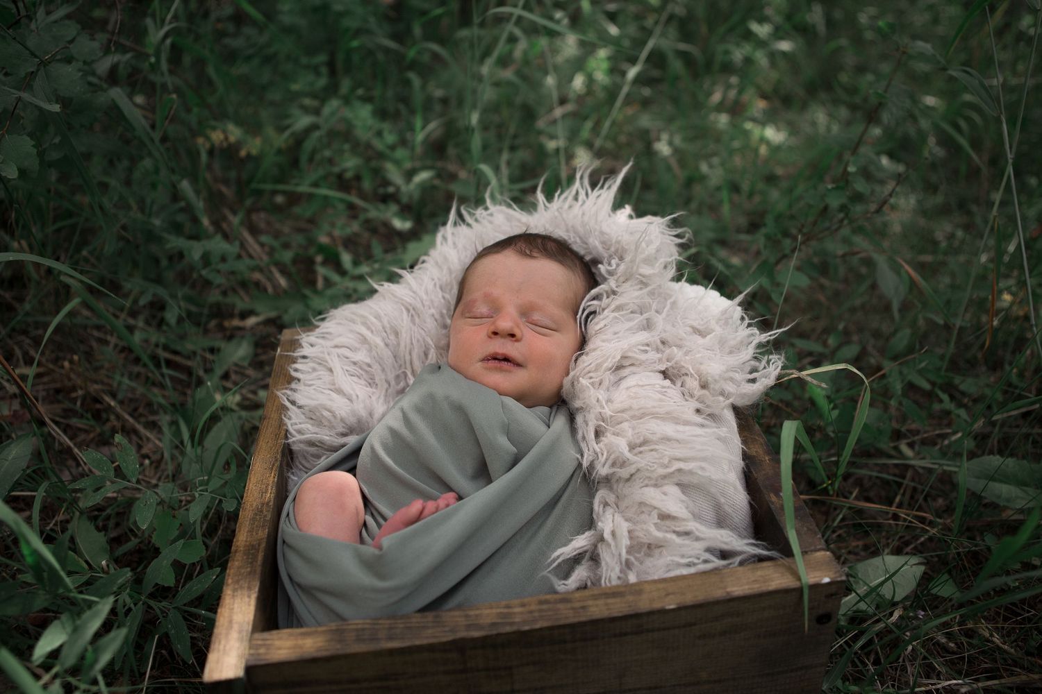 Sleeping baby wrapped in sage green fabric lies on white fur in wooden box amid green plants.