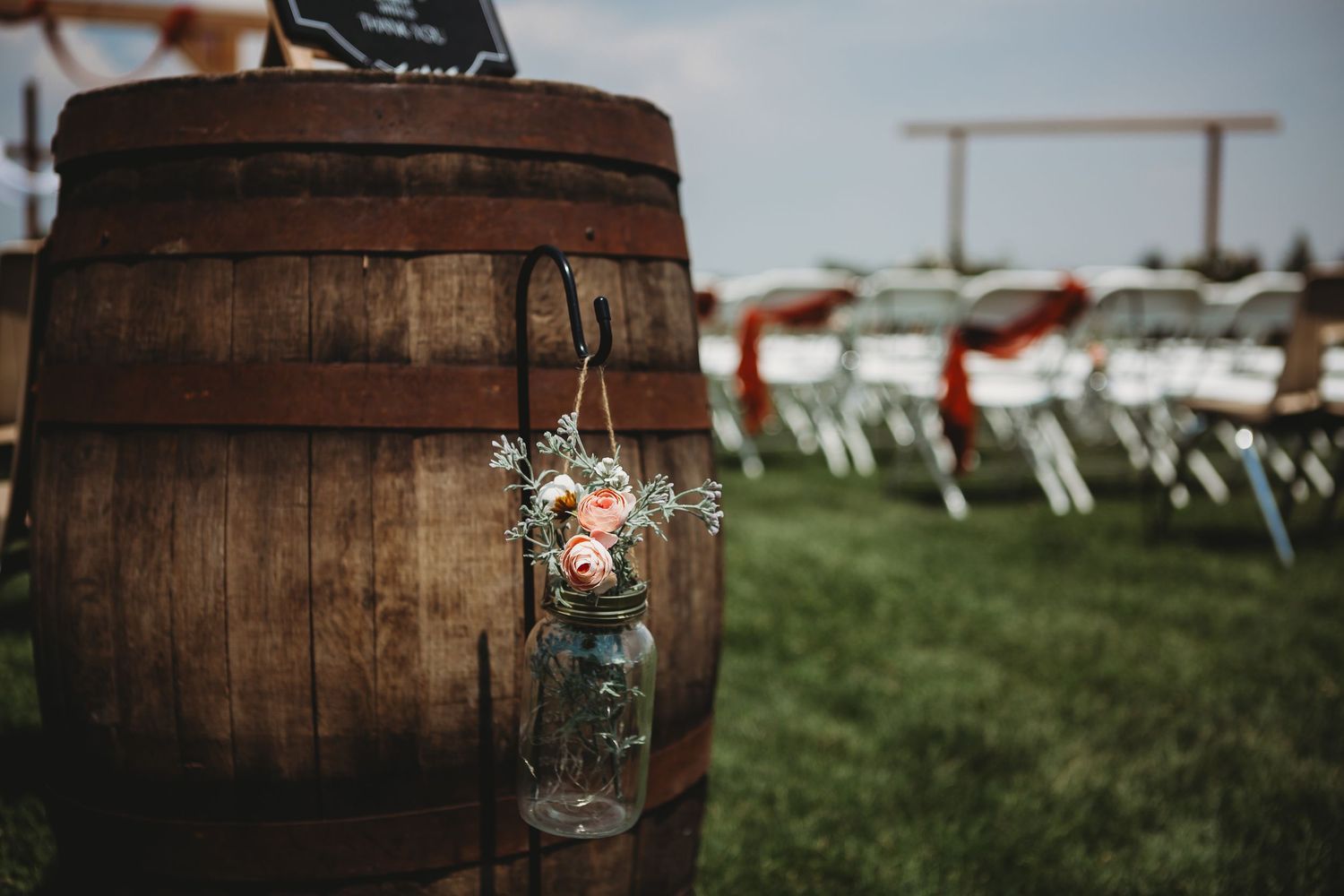 Rustic wedding decor featuring mason jar flower vases on wooden barrel with white chairs in background.