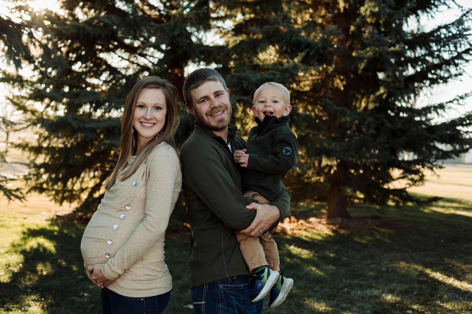 Maternity photo of couple in casual wear against pine trees at dusk.