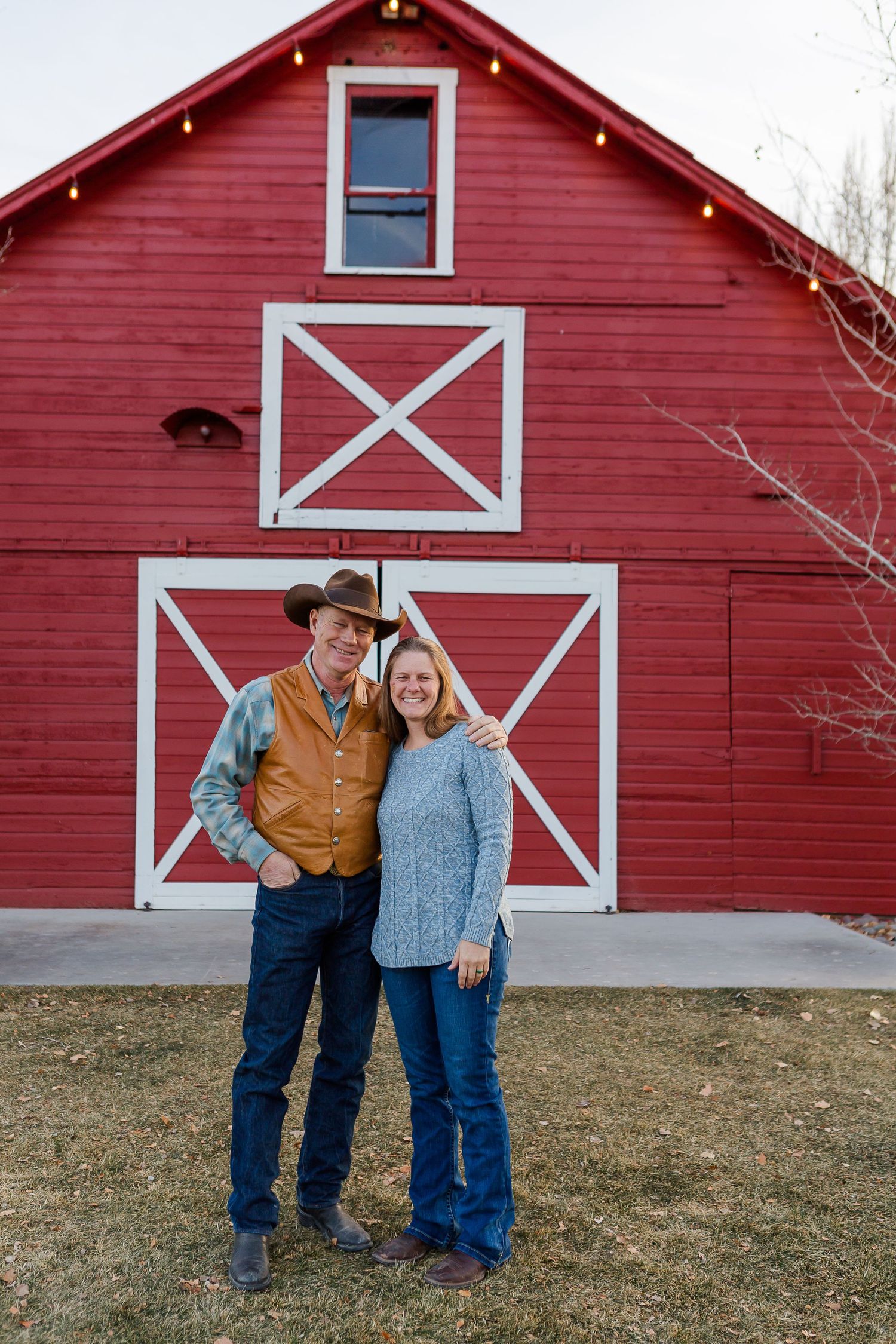Casual couple posing together against bright red barn with distinctive white X patterns.
