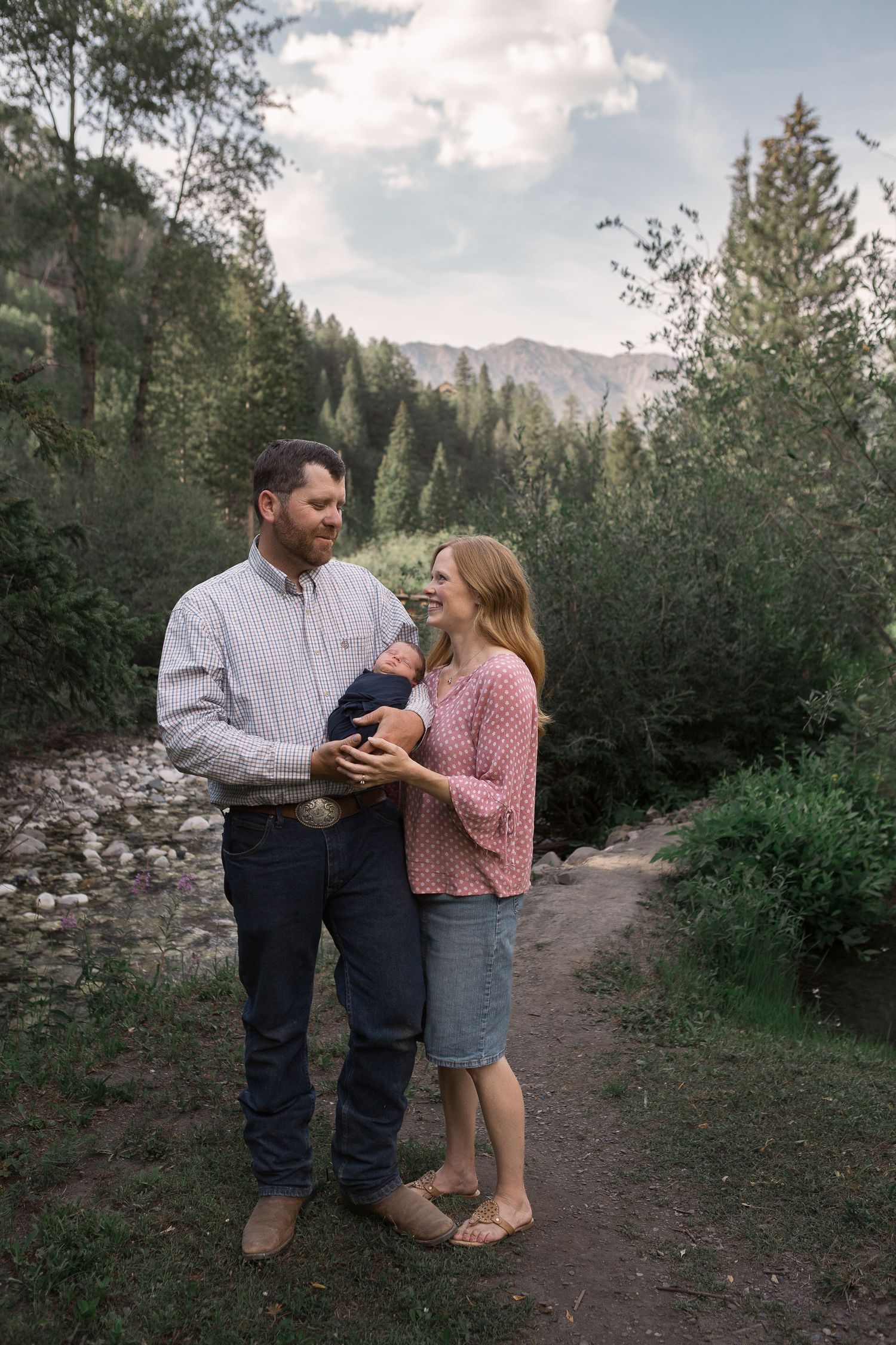 A couple stands together holding their newborn on a forested path with mountains in the background.