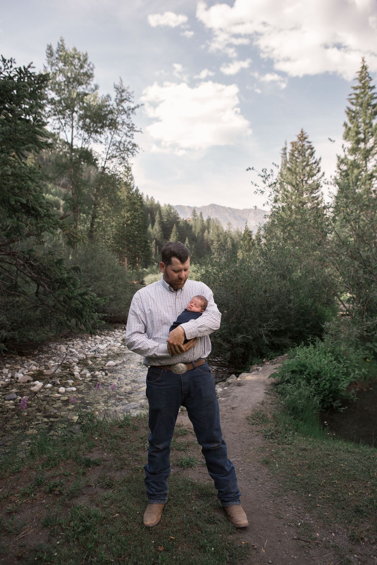 A person cradles a newborn baby while standing on a forest path with mountains in the background.