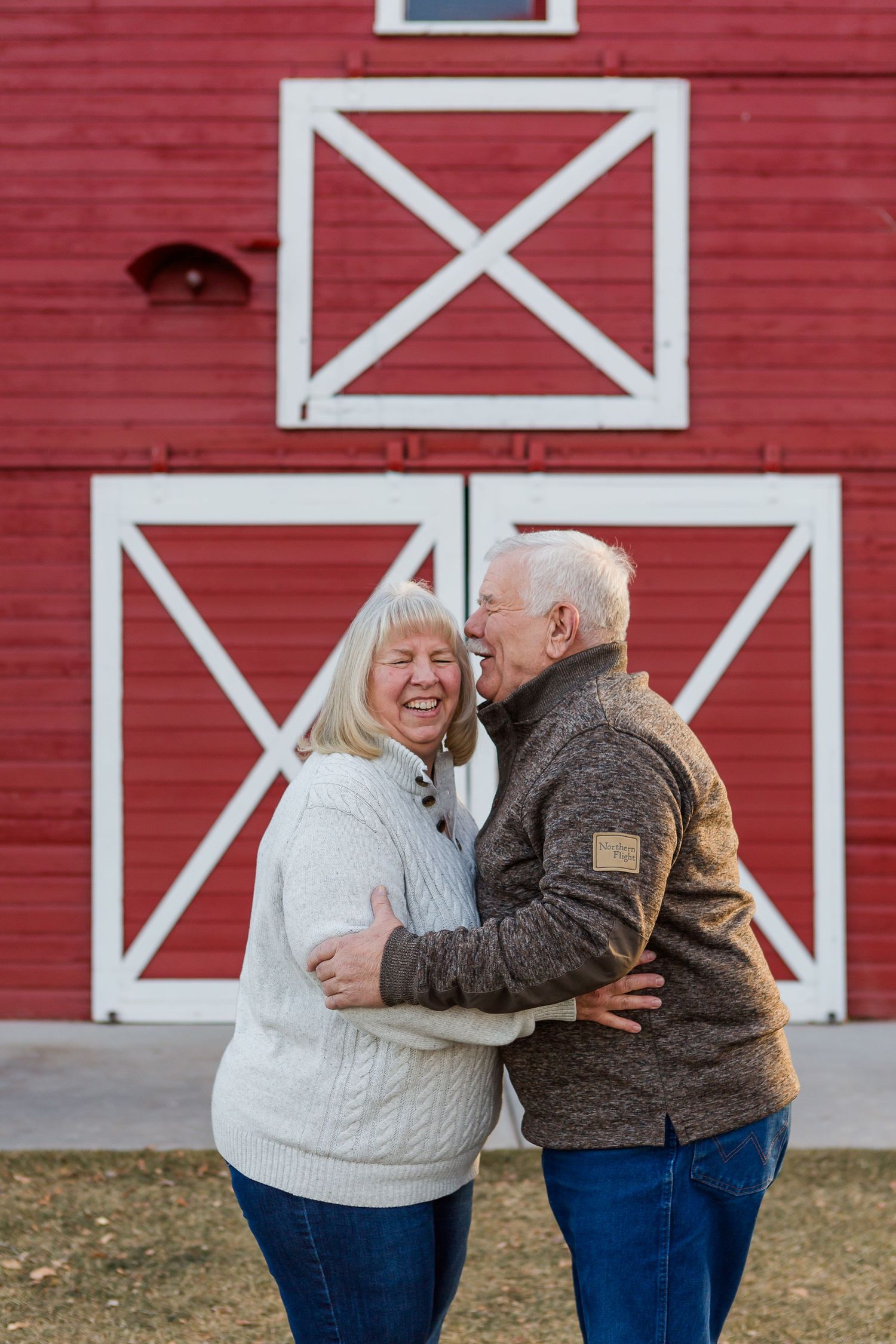 Couple embracing and laughing in front of a bright red barn with decorative white X designs.