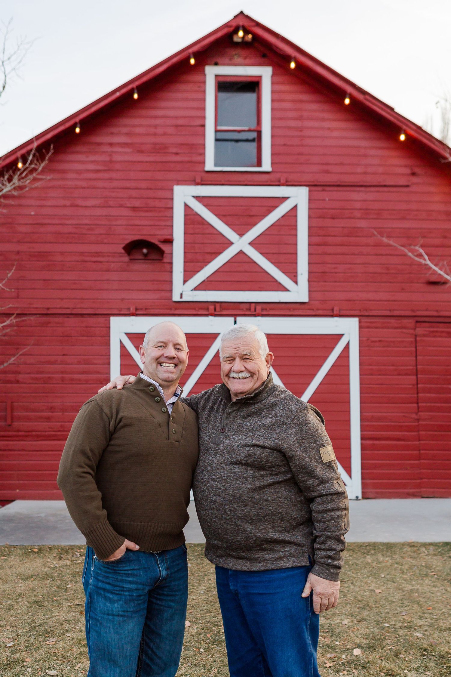 Two people smiling together in front of a red barn with white X patterns and holiday lights.