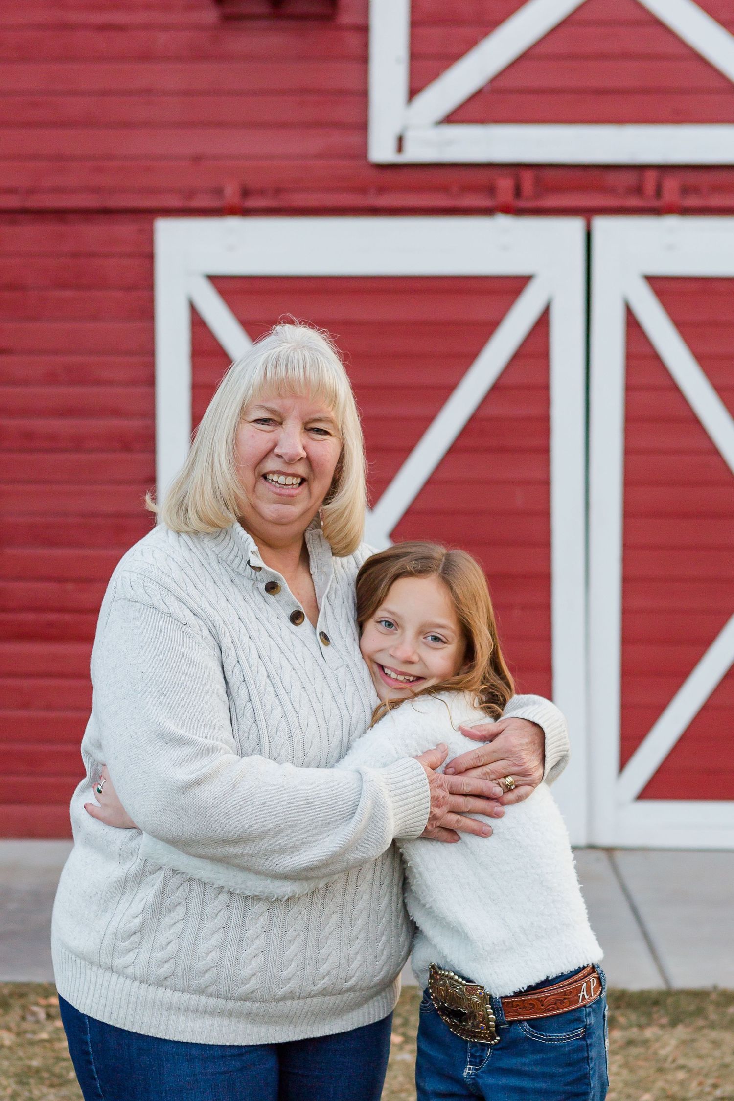 Two people in white sweaters embrace while standing in front of a bright red barn.