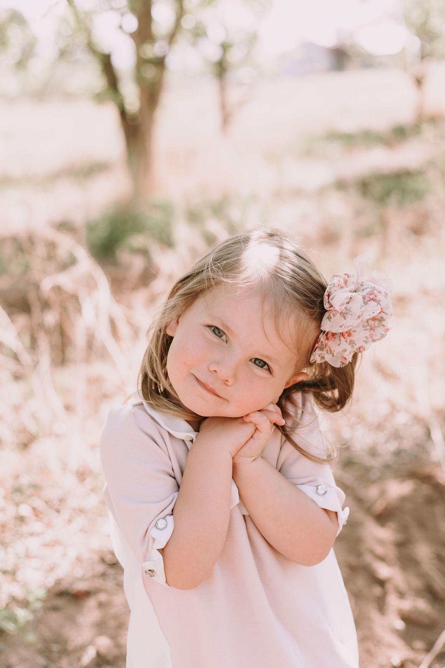 A child in a light colored dress poses with hands under chin while wearing a pink floral hair accessory in a dreamy outdoor setting.