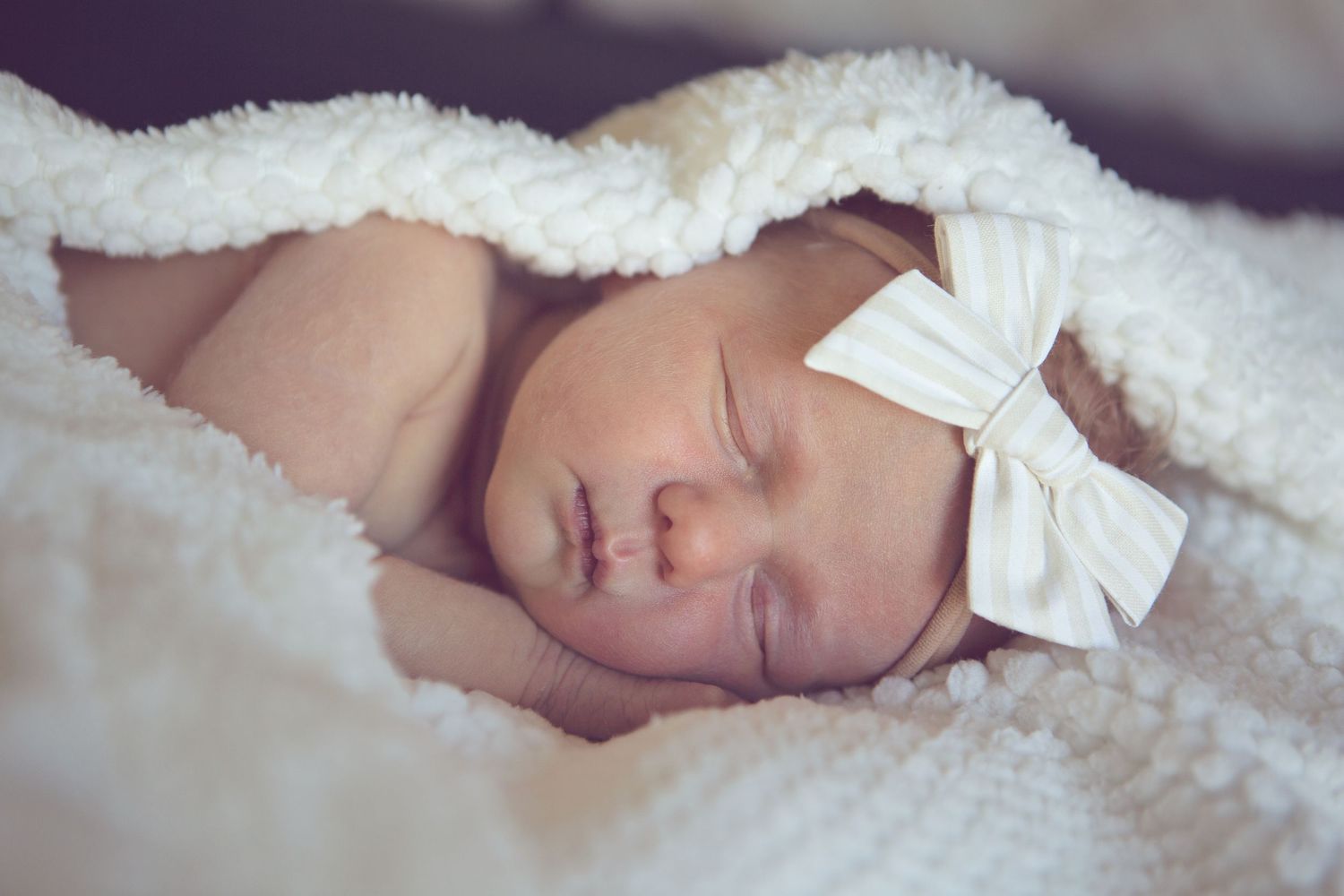Close-up of sleeping newborn with white bow headband wrapped in soft blanket.