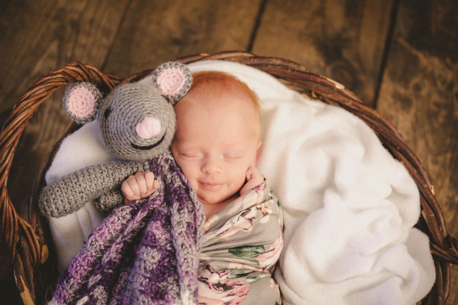 Sleeping newborn in basket with knit mouse toy and soft white blanket during professional photoshoot.
