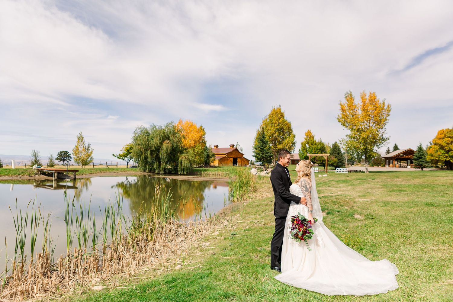 A wedding couple stands by a serene lake with fall trees and a rustic building in the background.