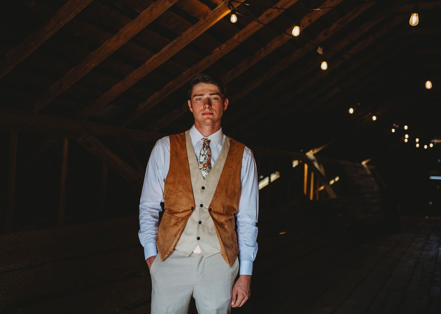 Groomsman poses in a rustic barn wearing a tan vest and gray suit.
