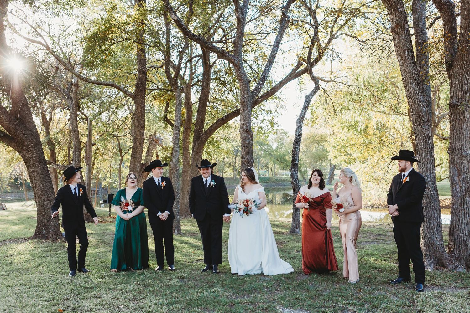 A wedding party poses together under tall trees in a park during golden hour.