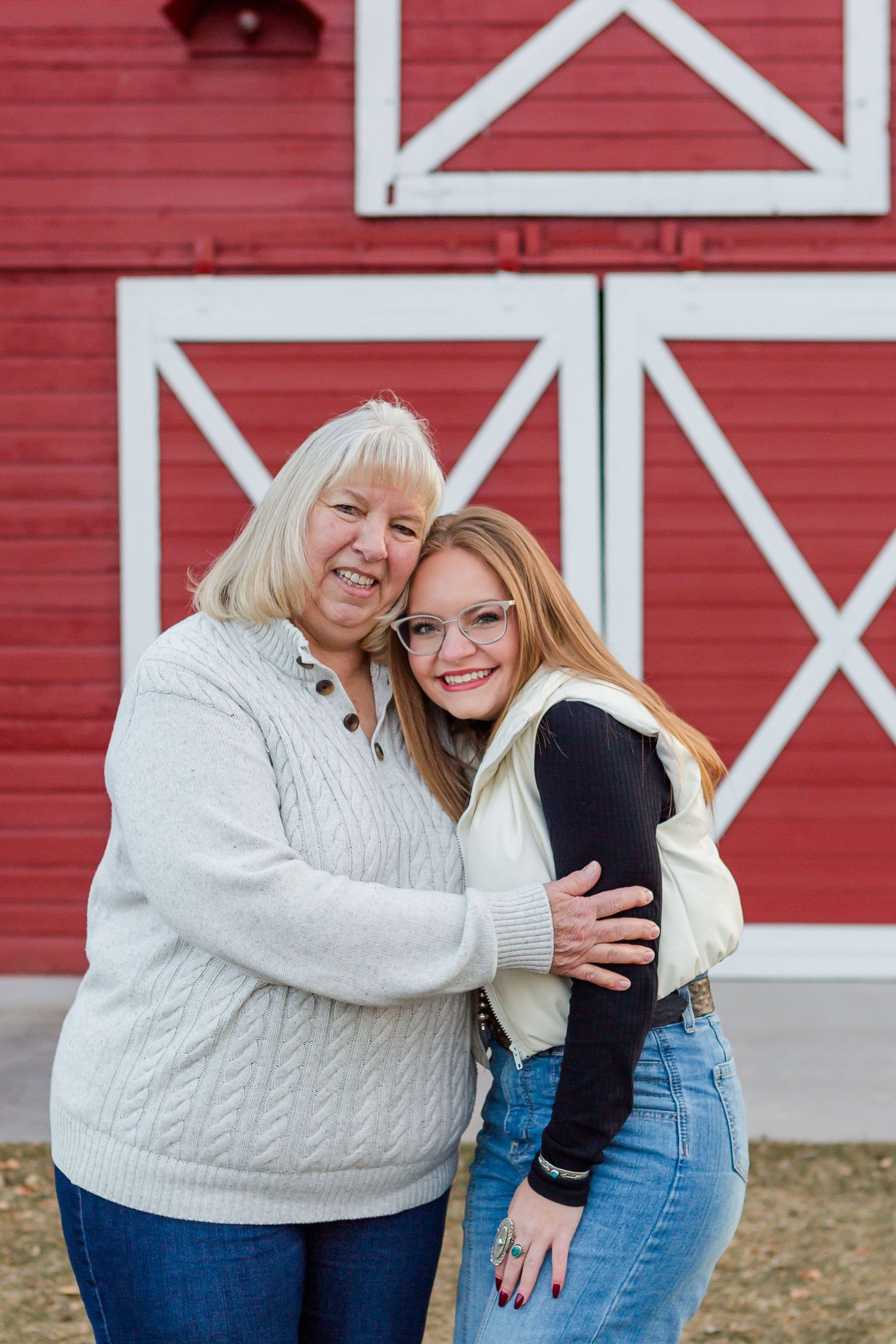 Two people share a warm embrace while standing in front of a red barn with white X patterns.