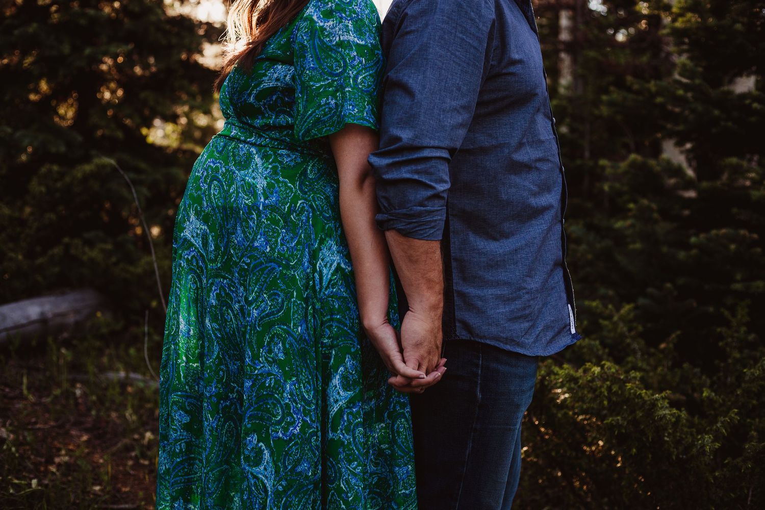 A couple holding hands while standing together in a forest, wearing a teal patterned dress and navy shirt.