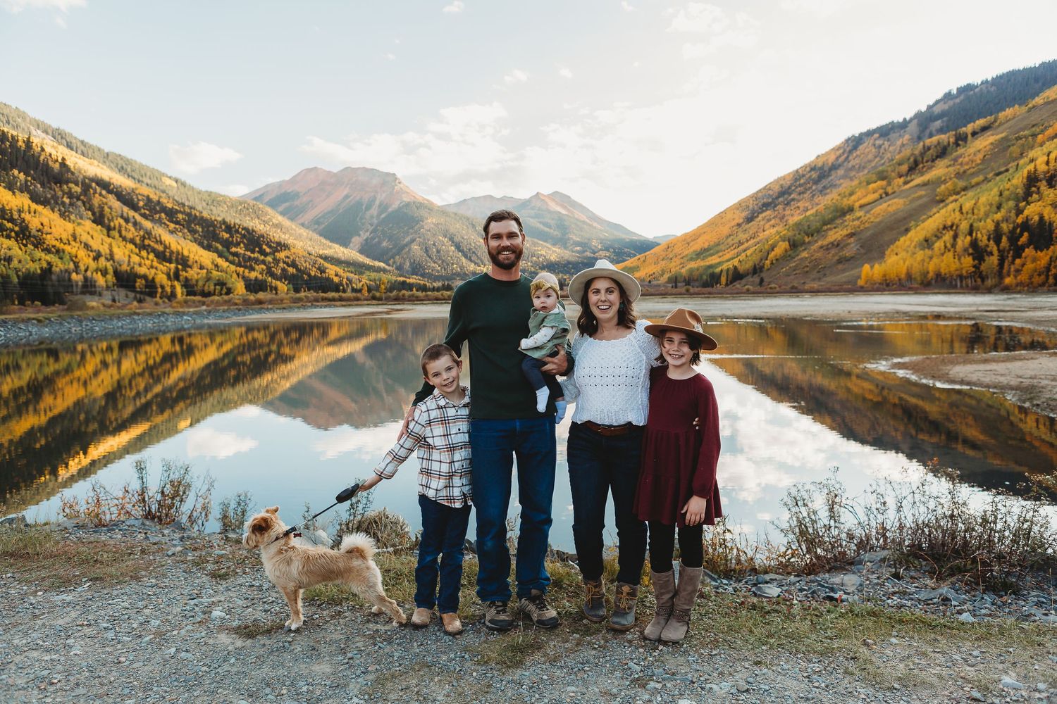 Group stands with dog by mountain lake with golden autumn trees and perfect reflections in calm water.