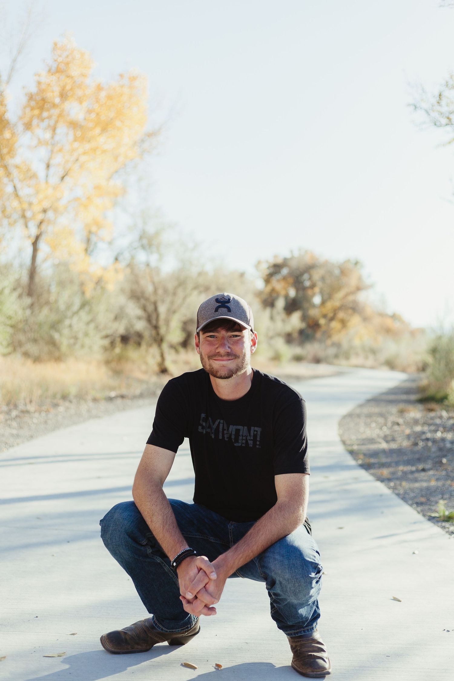 Person in baseball cap crouching on concrete path with autumn foliage in background.