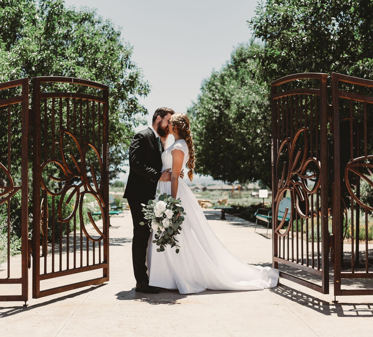 A romantic wedding moment between ornate rustic metal gates in a garden setting.