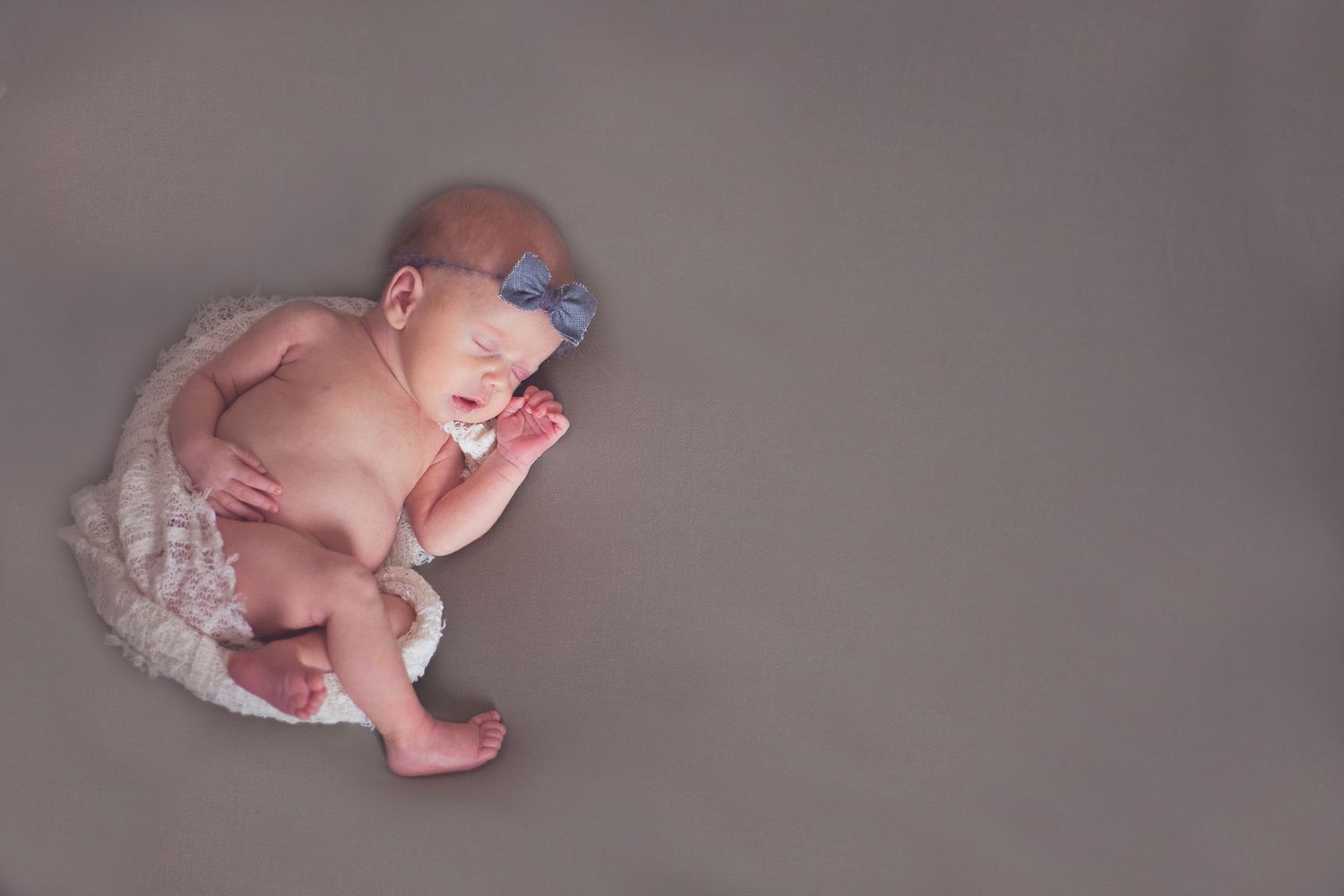 Sleeping newborn in tulle skirt and blue bow positioned on grey backdrop.