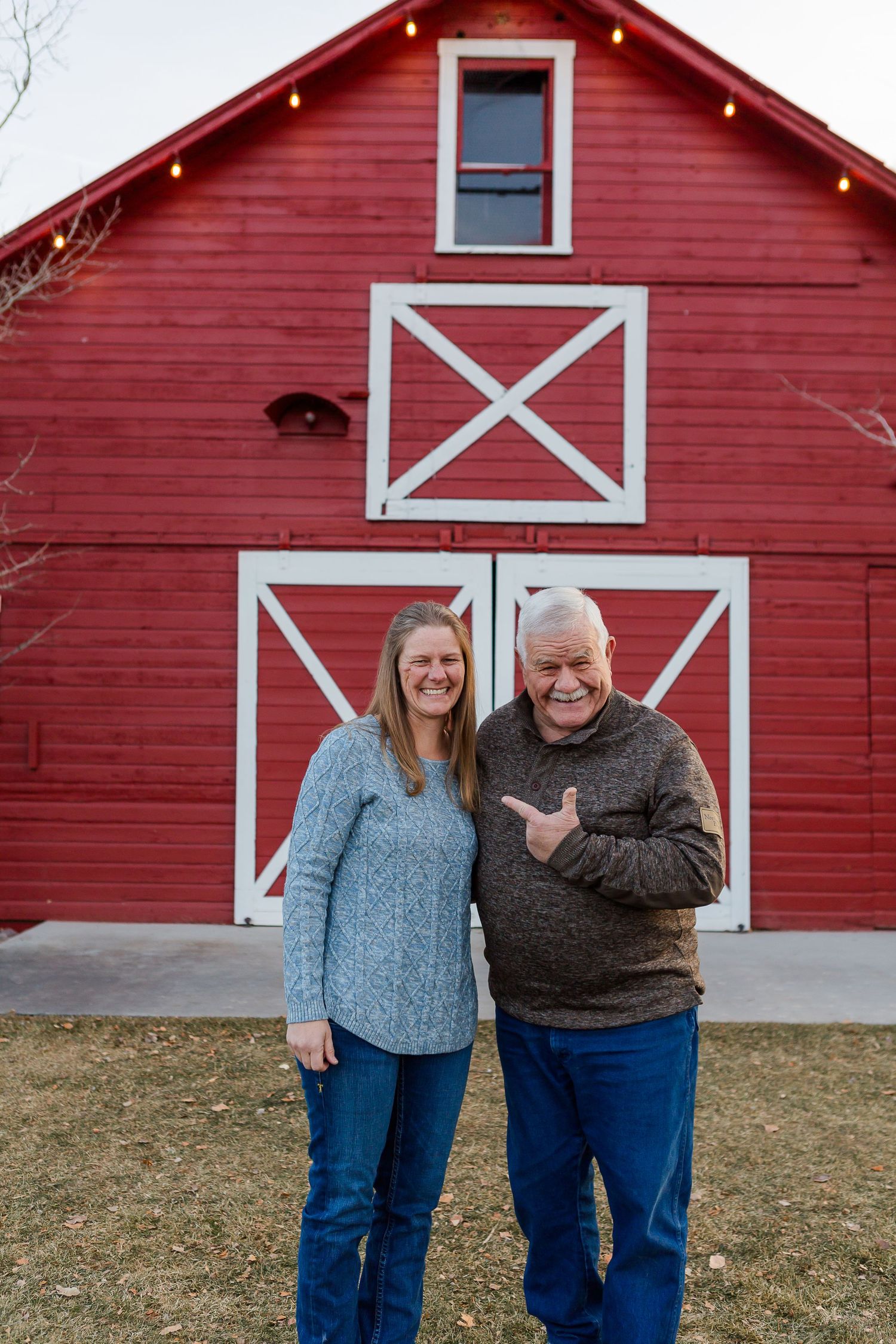 Two people posing together in front of a red barn with decorative white X patterns and string lights.