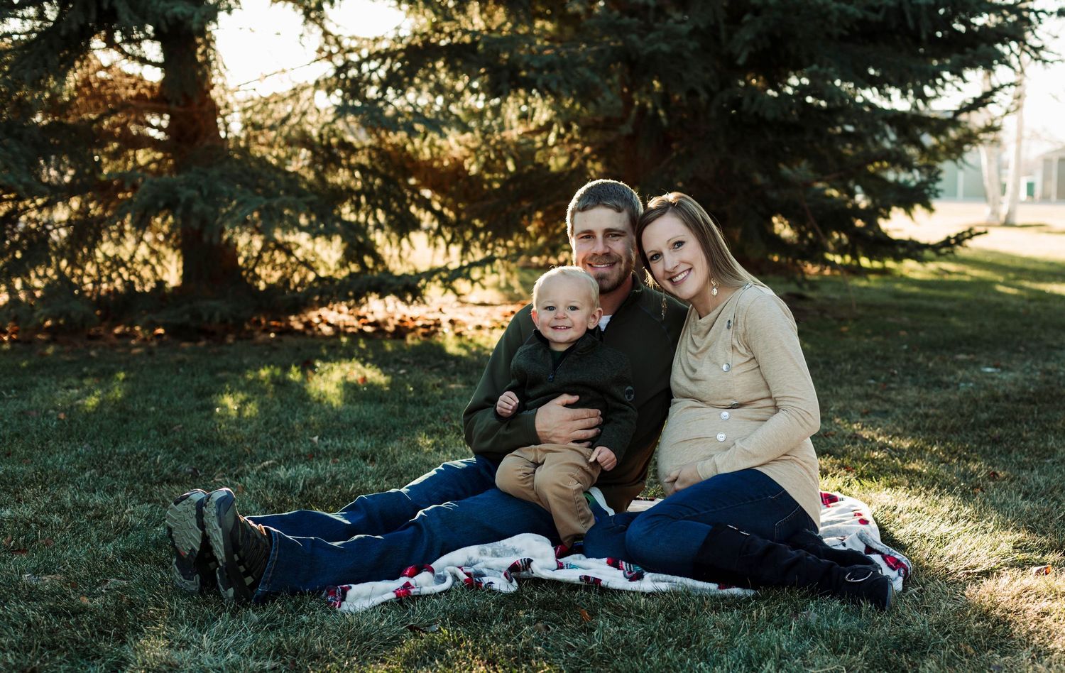 Family relaxes on blanket during outdoor maternity photography session near evergreen trees.