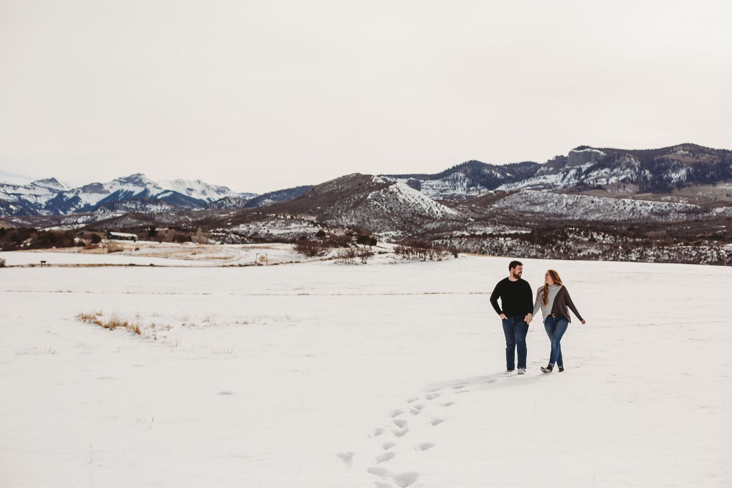 Two people walking through a vast snowy landscape with mountains in the distance.