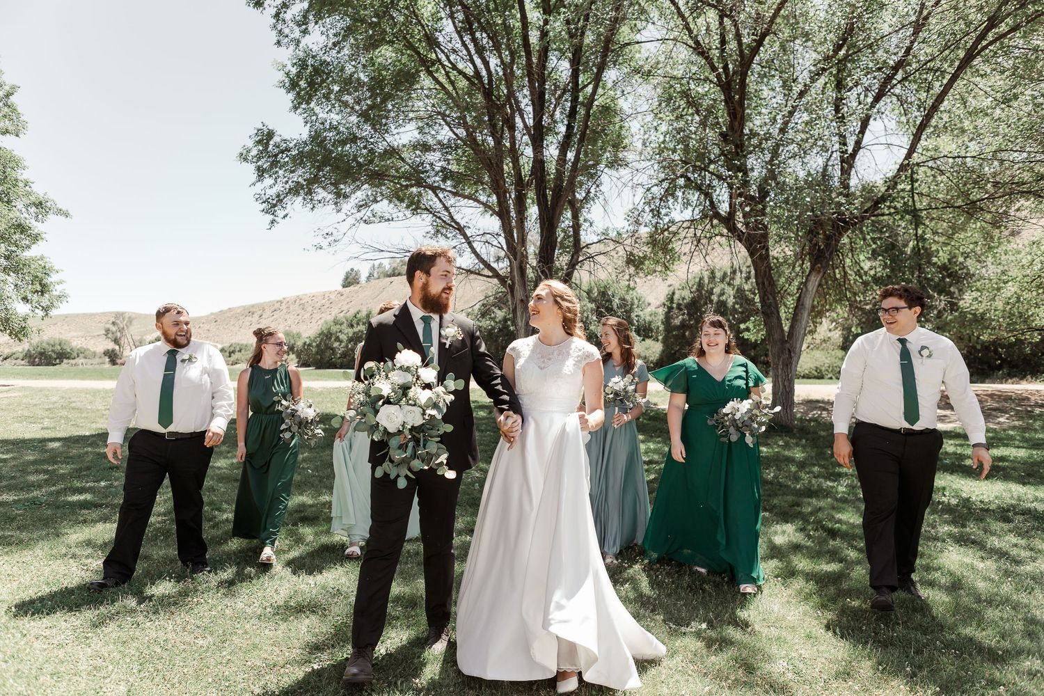 Outdoor wedding party photo with bridesmaids in emerald and mint dresses under trees.