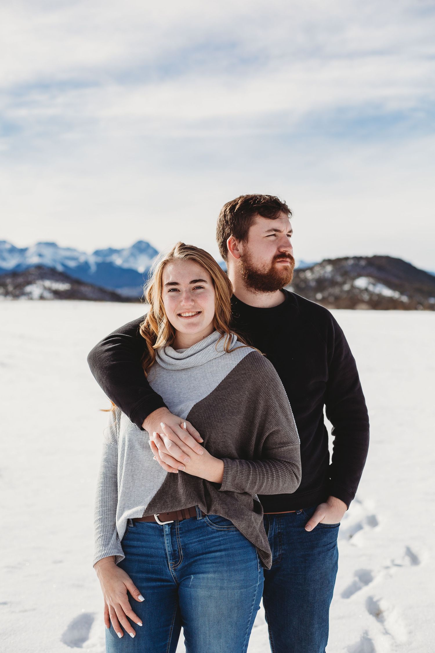 A couple poses together in winter attire against a snowy mountain landscape.