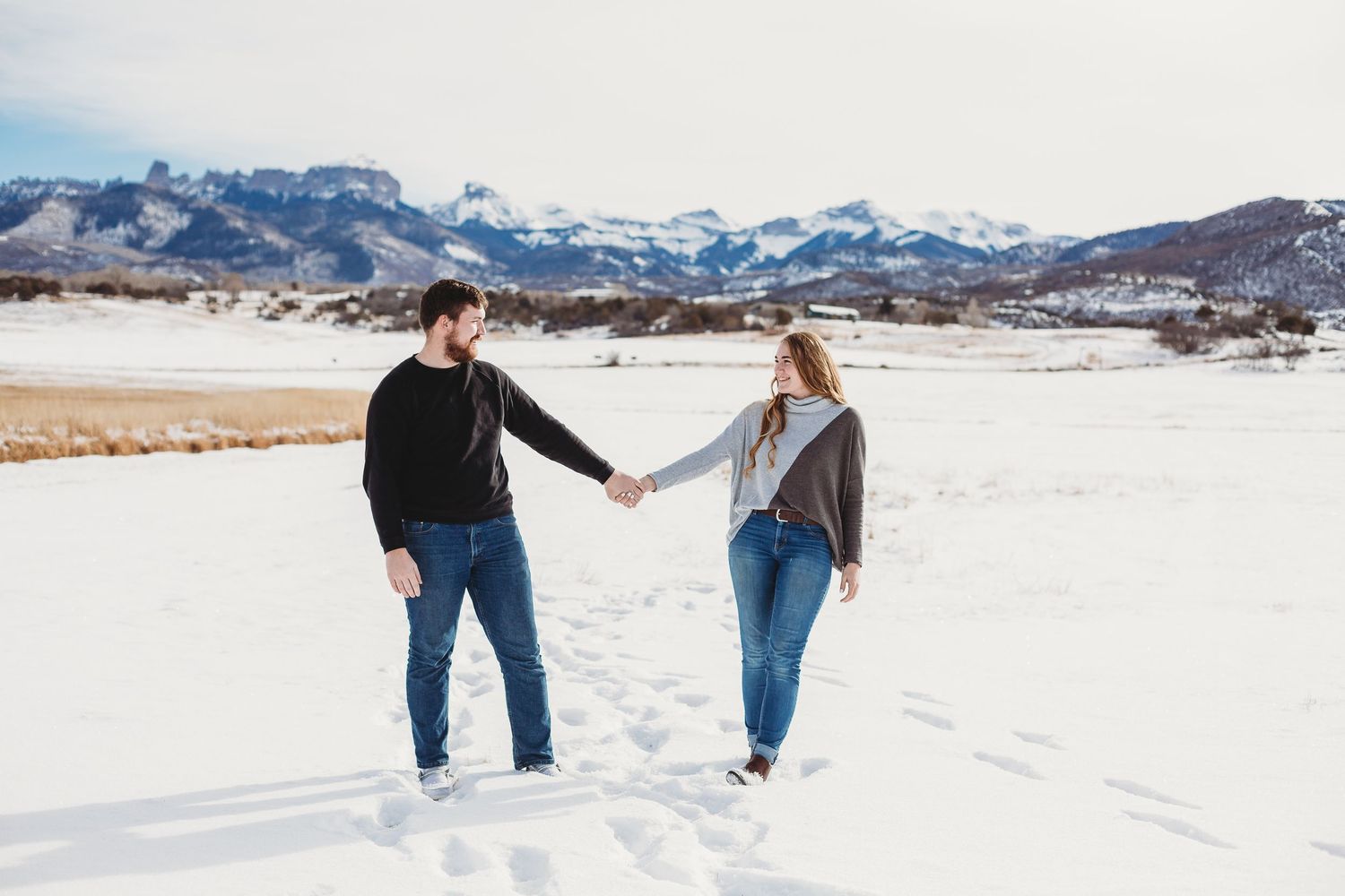 A couple holds hands while walking together on a snowy field with mountain ranges in the background.