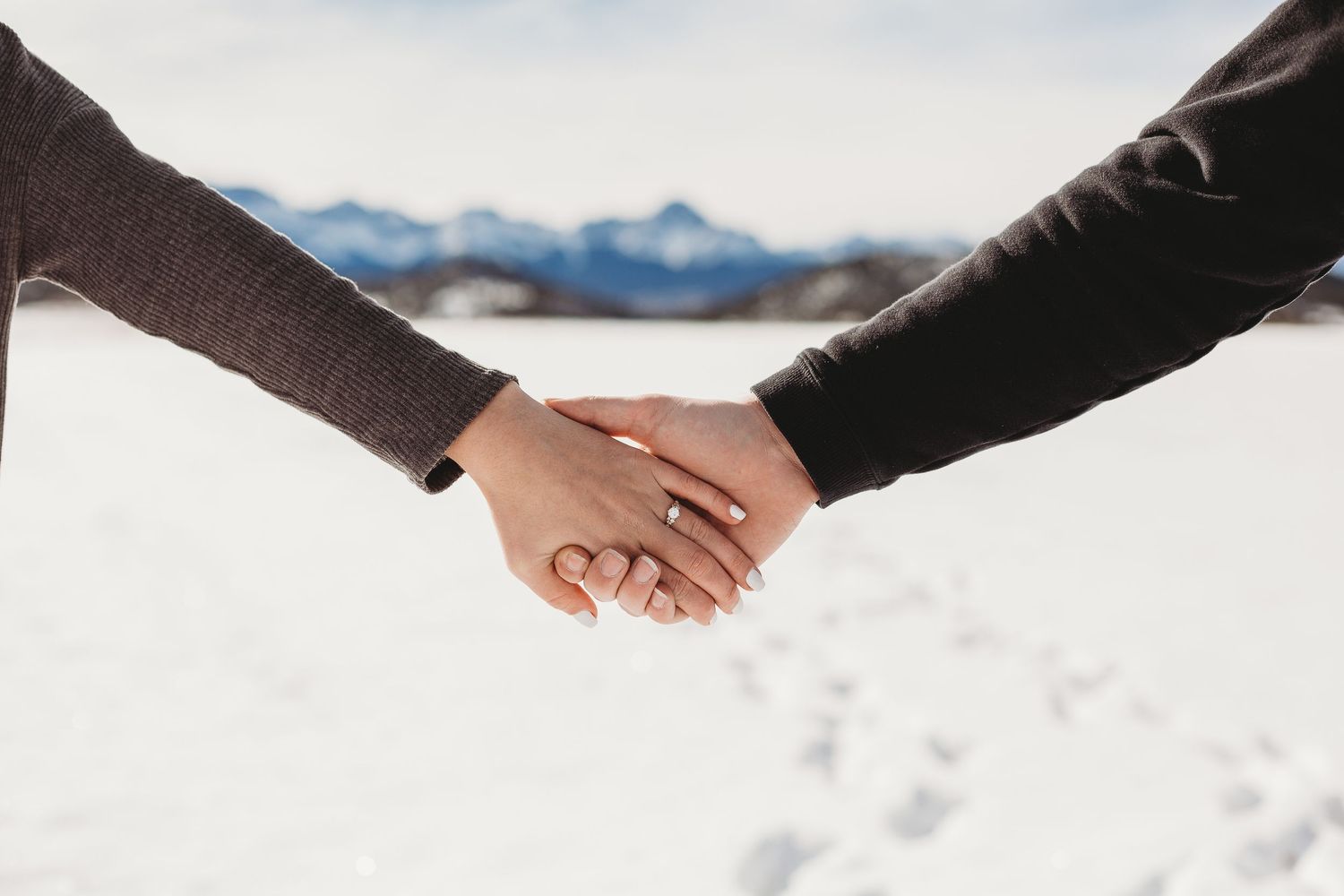 Two people holding hands with snow-covered mountains and footprints visible in the background.