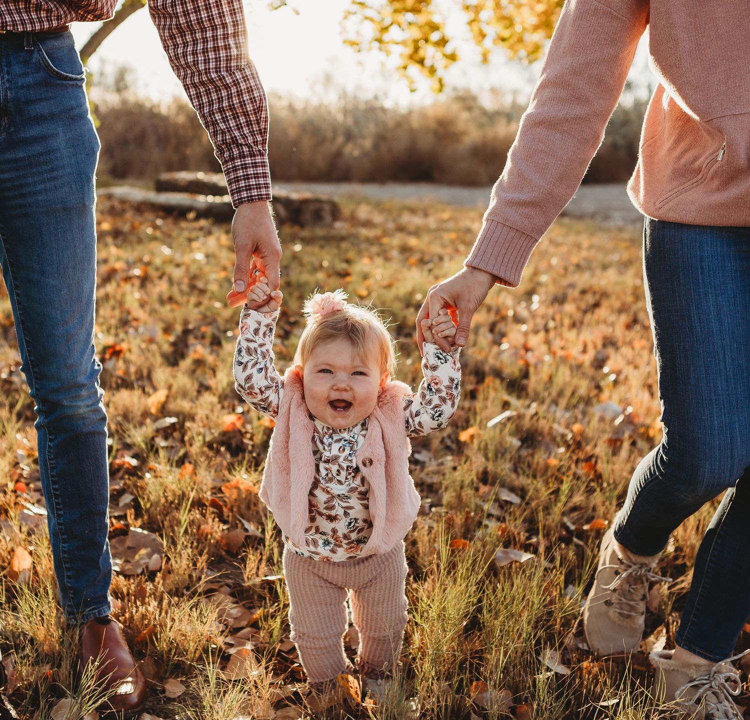A happy toddler walks while holding parents' hands in a sunlit autumn field.