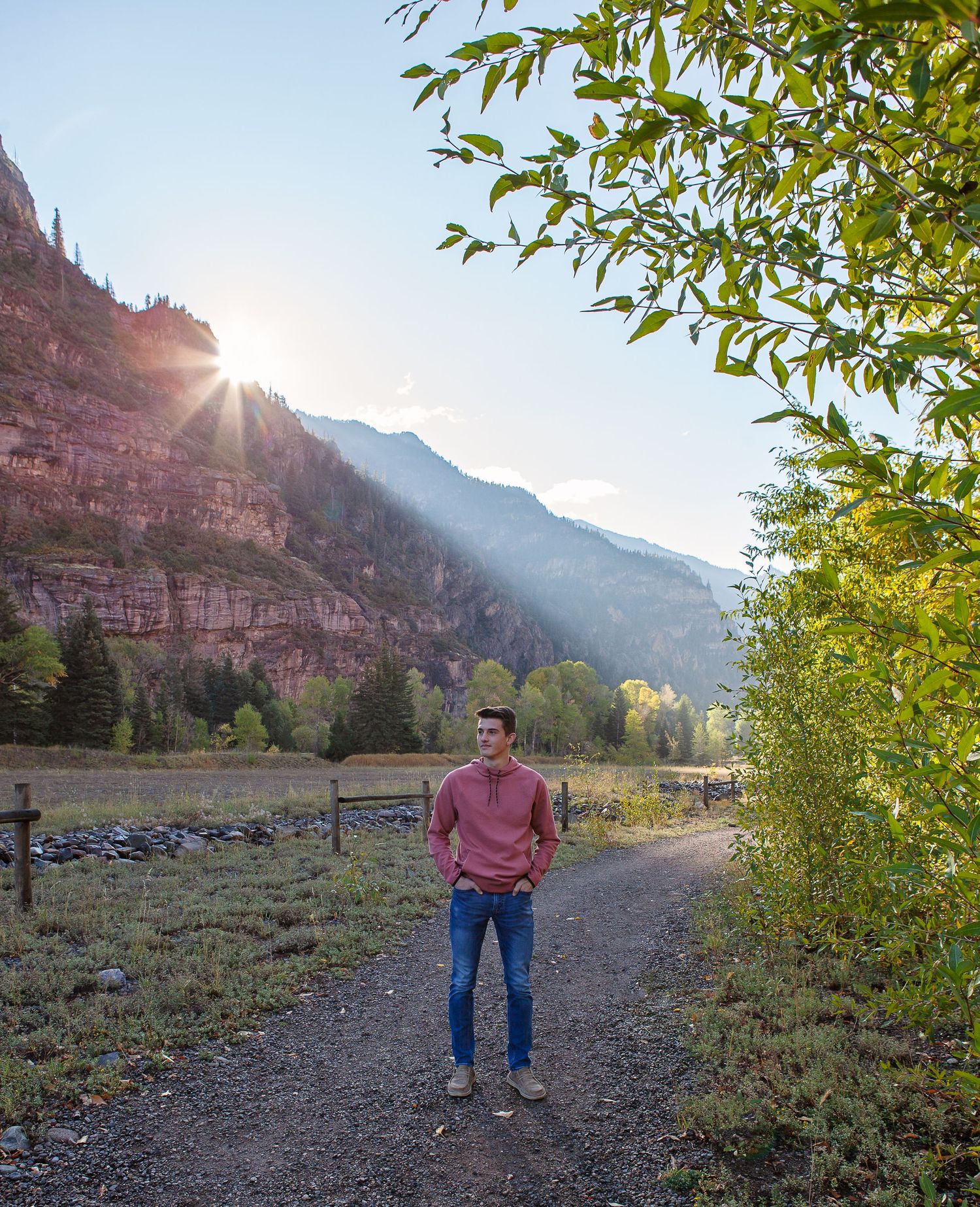 A person poses on dirt path with dramatic mountain landscape and sunburst behind.