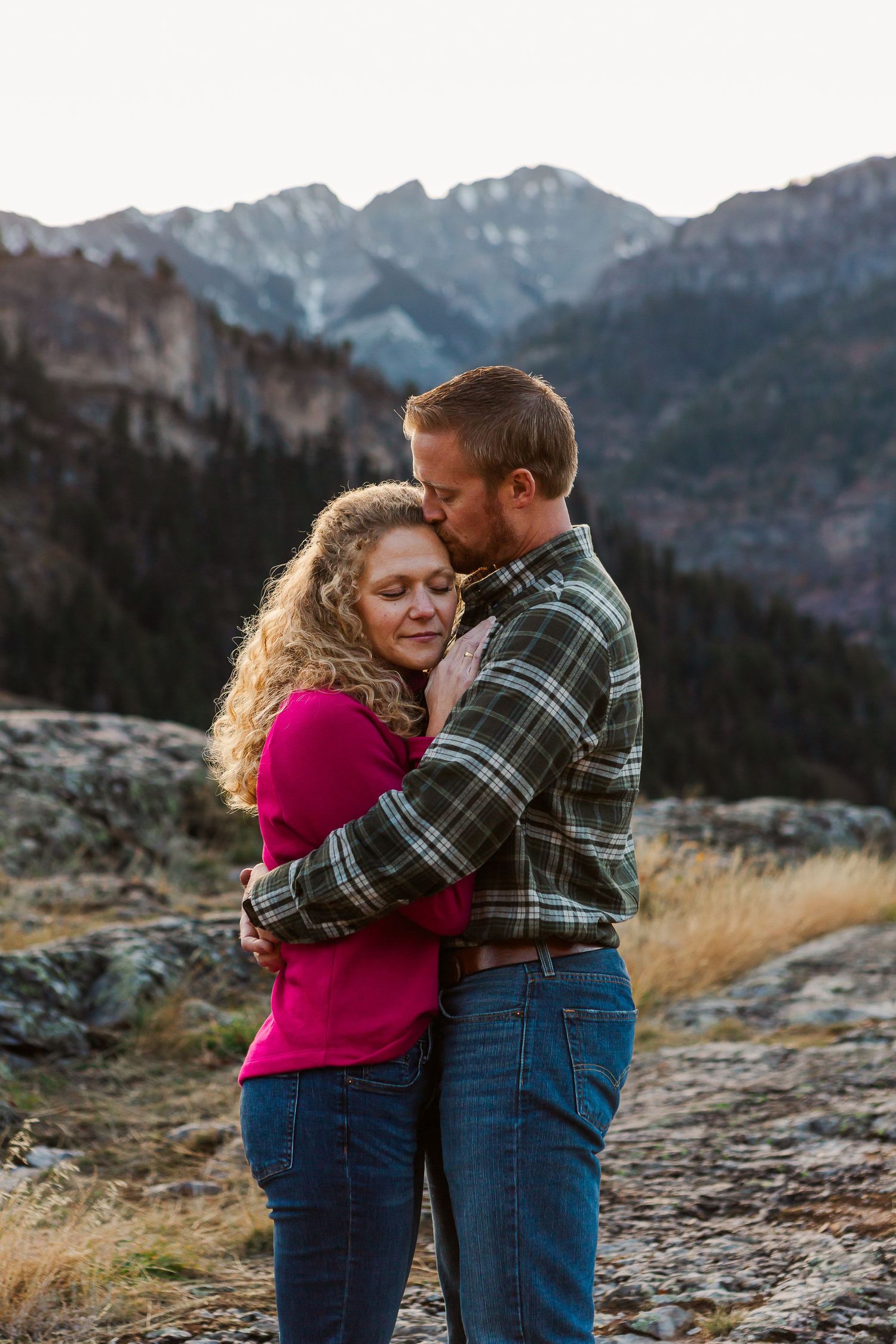 A couple embraces while standing on rocky terrain with snow-capped mountains in the background at sunset.