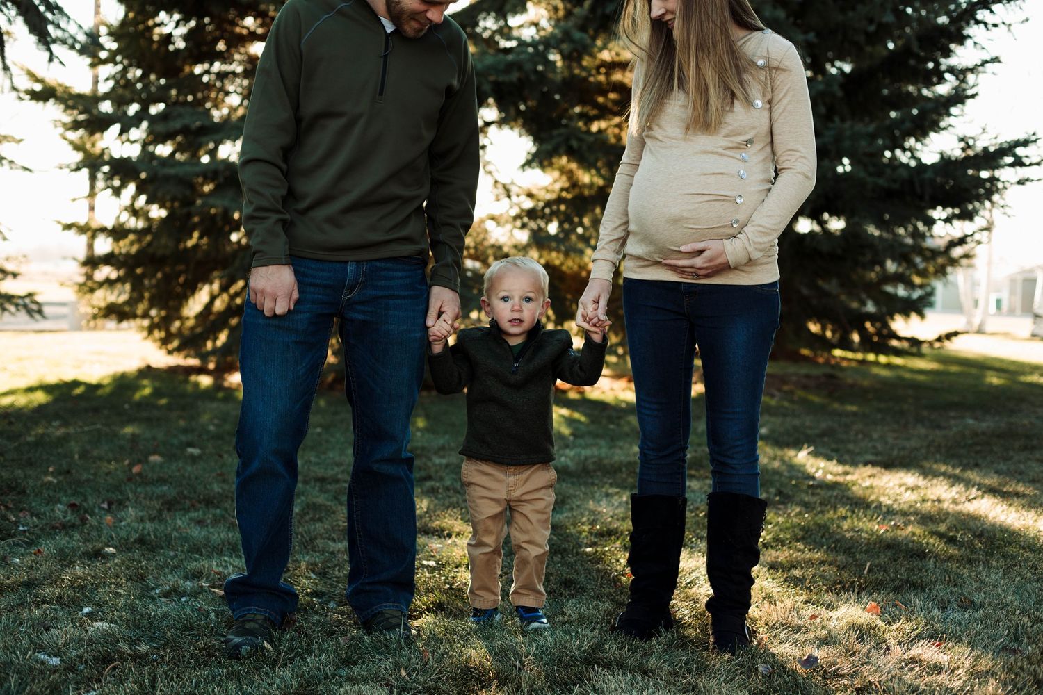 Family holds hands with young child while standing in front of pine trees at sunset.