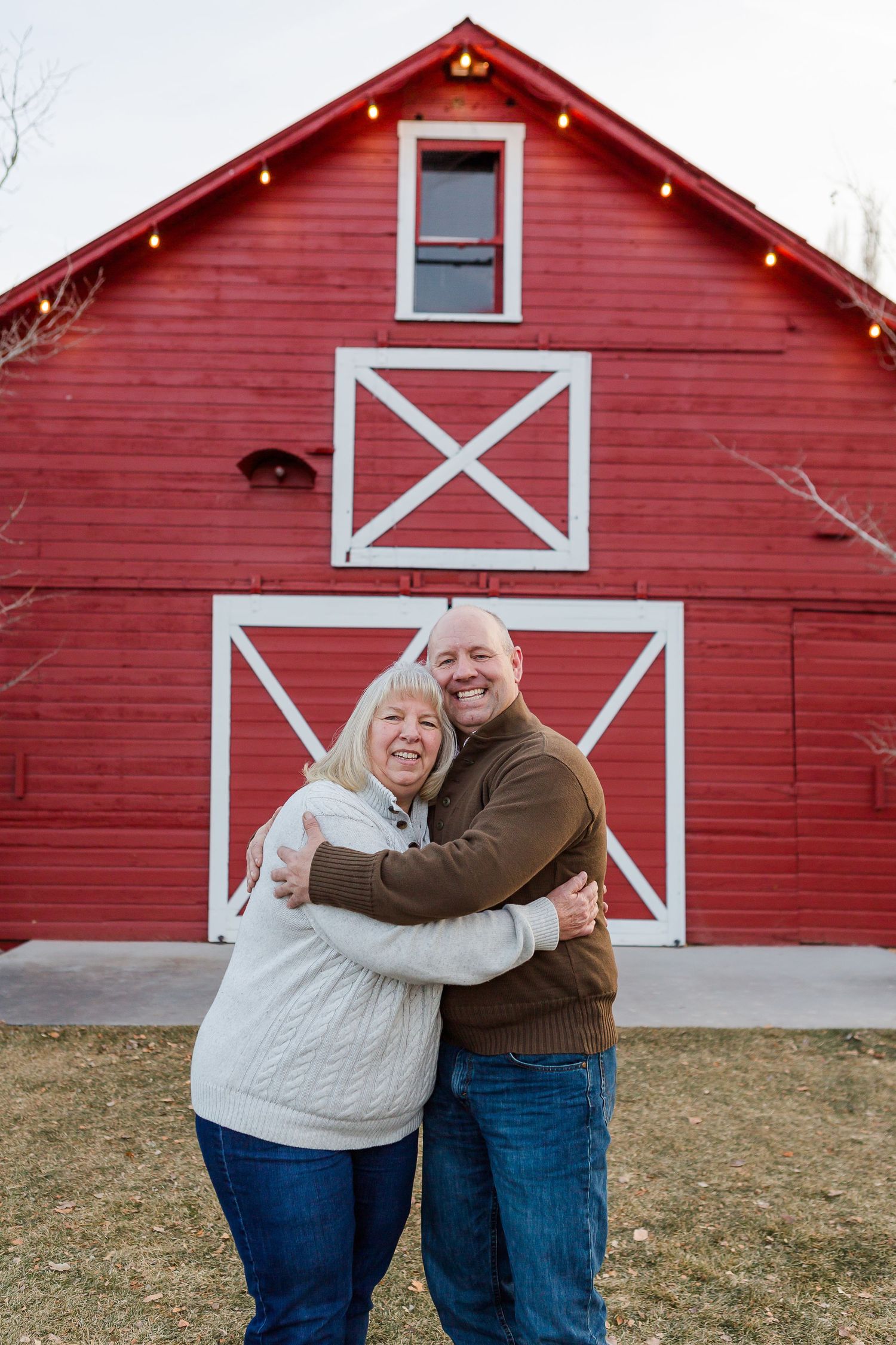 Two people share a joyful embrace in front of a bright red barn with decorative white trim and lights.