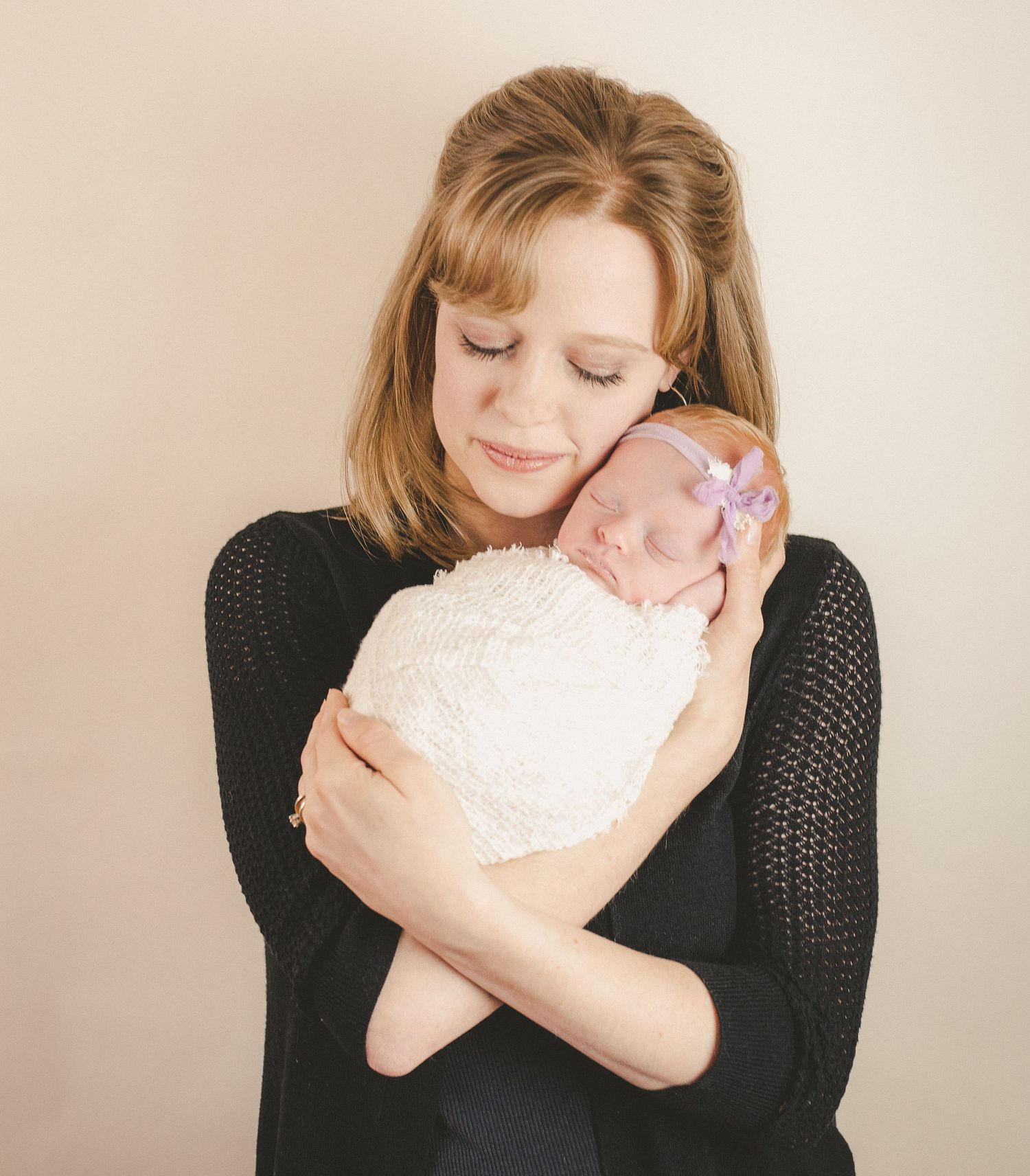Tender moment captured as woman in black sweater cradles sleeping newborn wrapped in white.