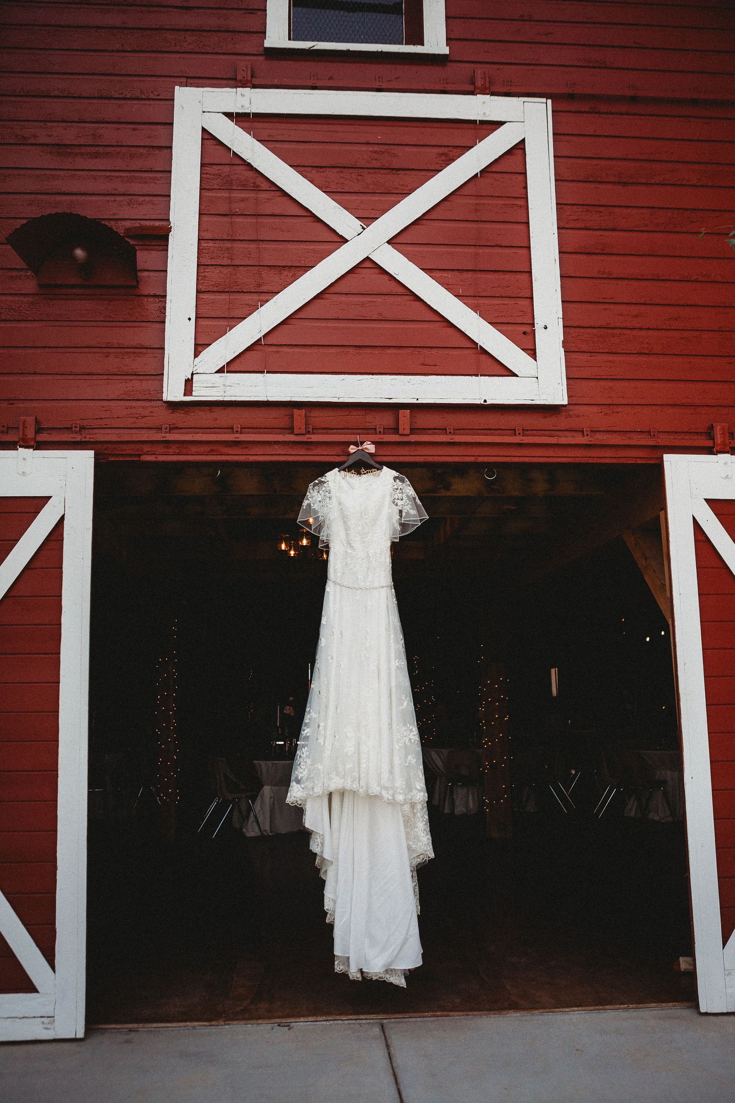 A delicate white lace wedding dress hangs from barn doors on a rustic red building.