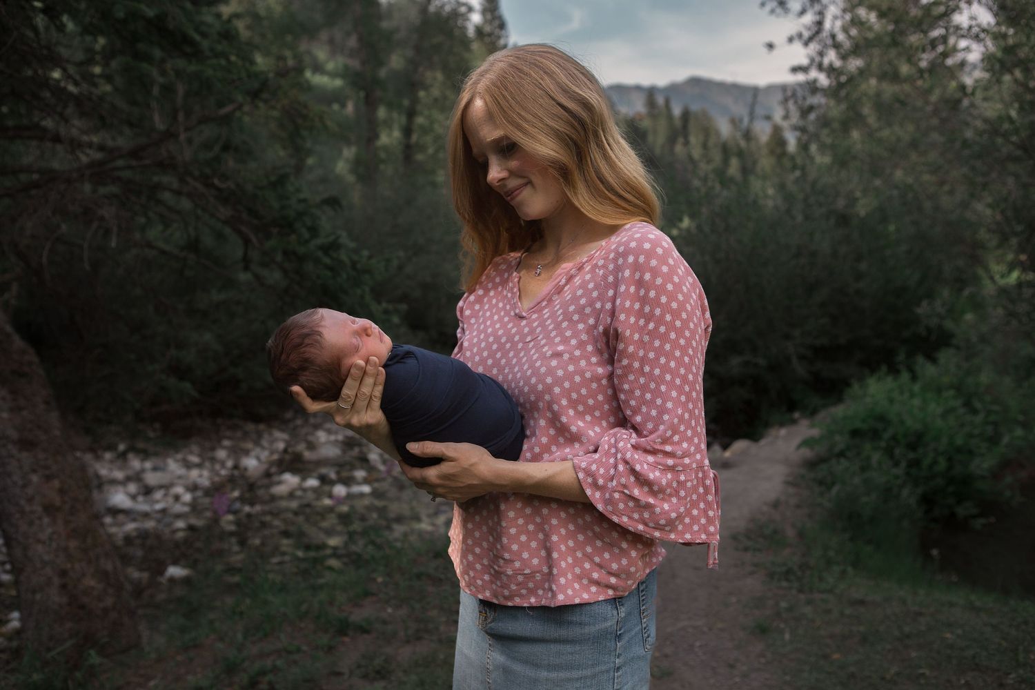 Pink-clothed figure cradles infant on nature path with mountain stream backdrop.