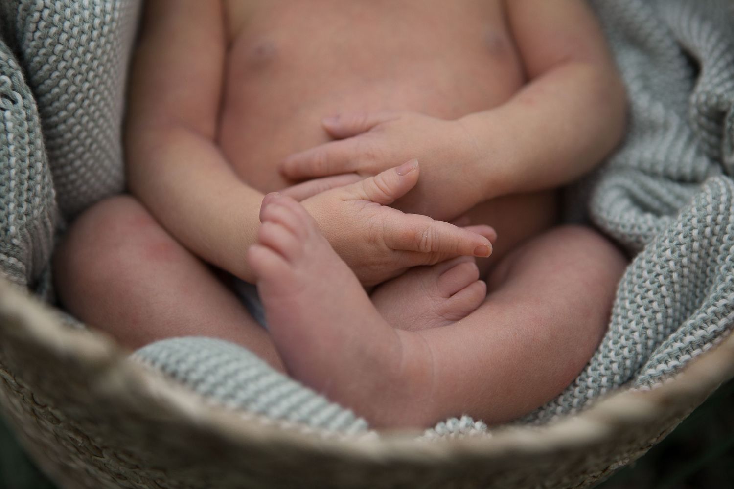 Close-up detail of tiny baby hands and feet with gray knit blanket in basket.