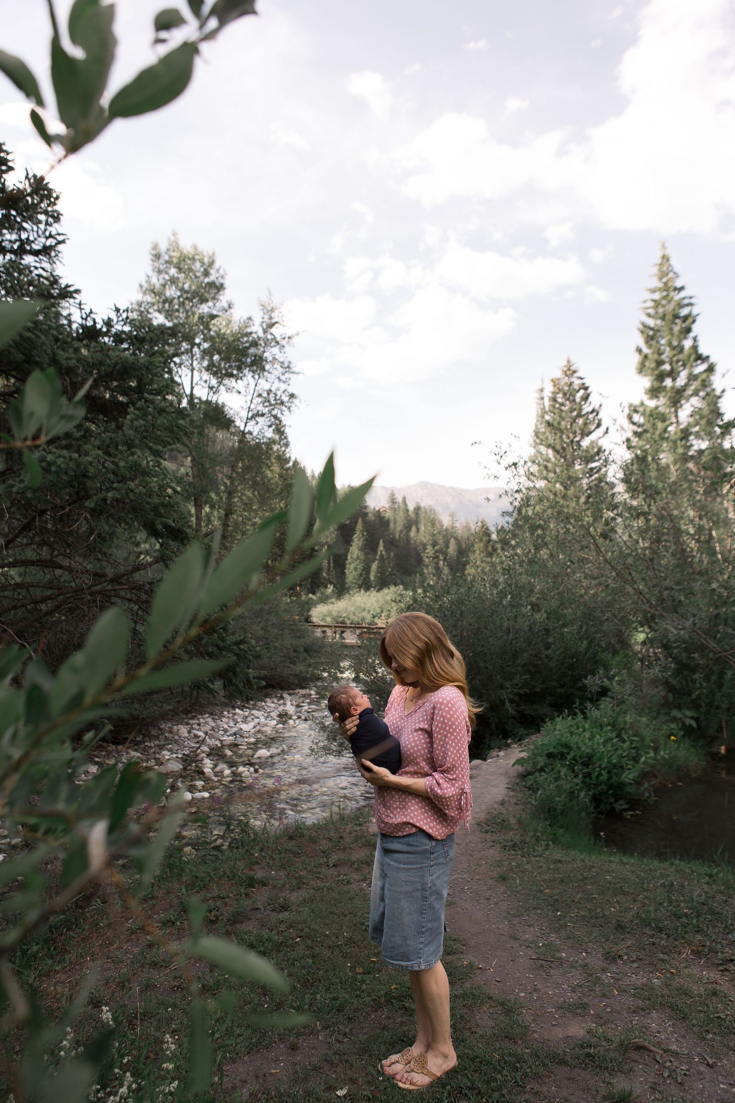 A person stands holding an infant along a scenic mountain trail by a flowing stream.