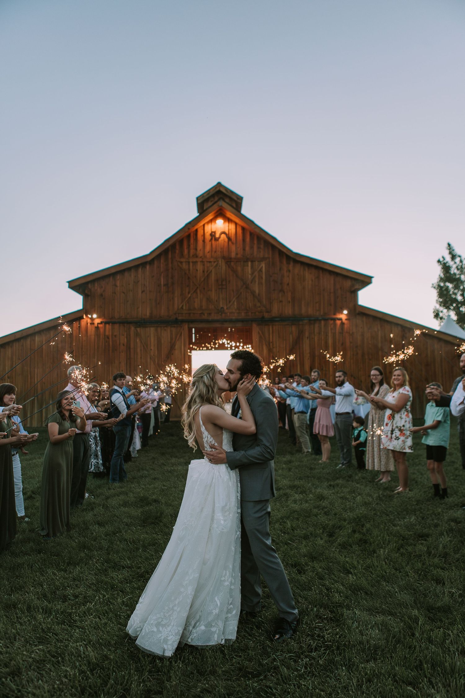 Evening outdoor first dance with guests surrounding couple outside rustic barn venue at sunset.