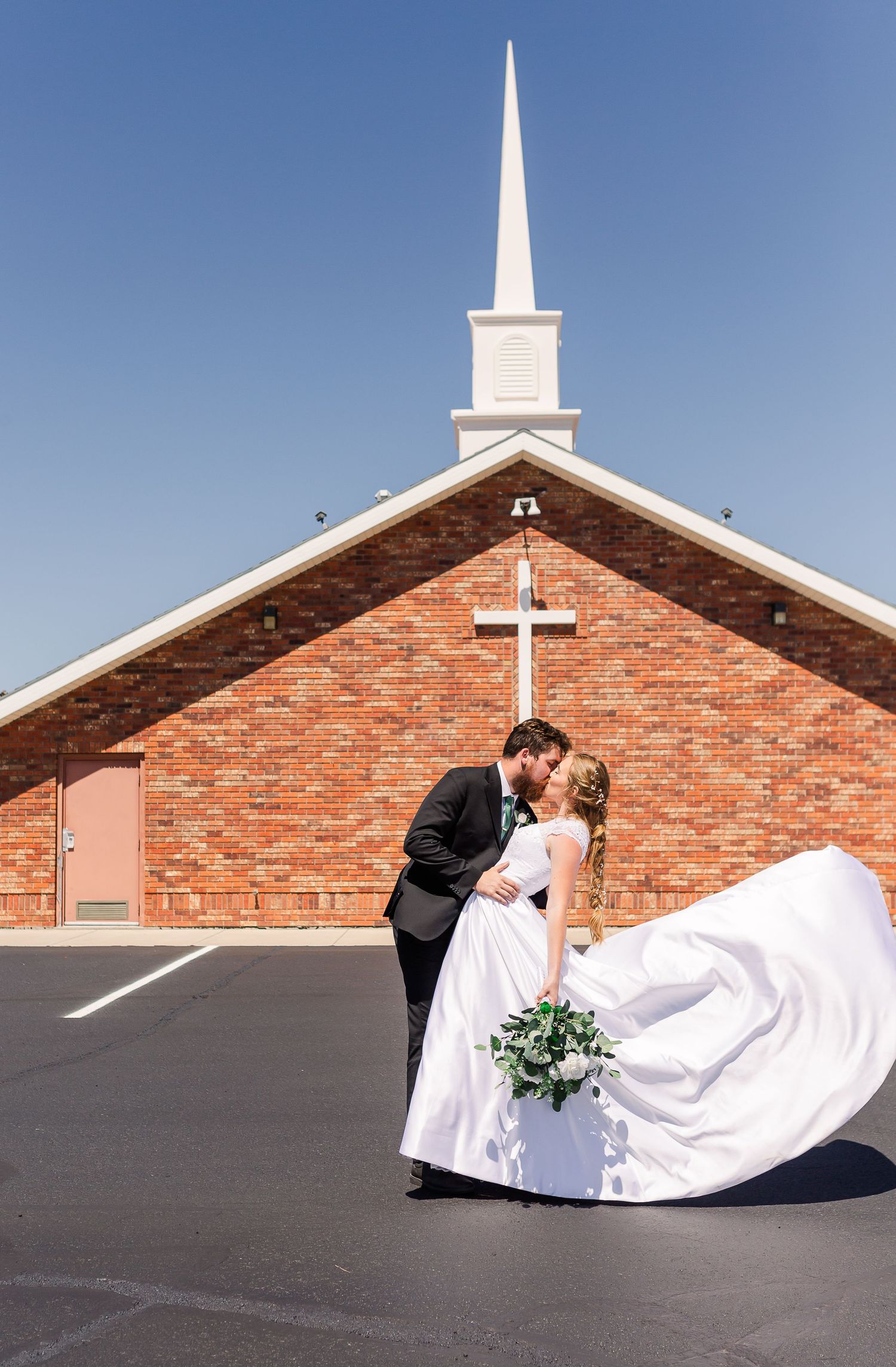 A romantic wedding photo outside a brick church with a white steeple against a bright blue sky.