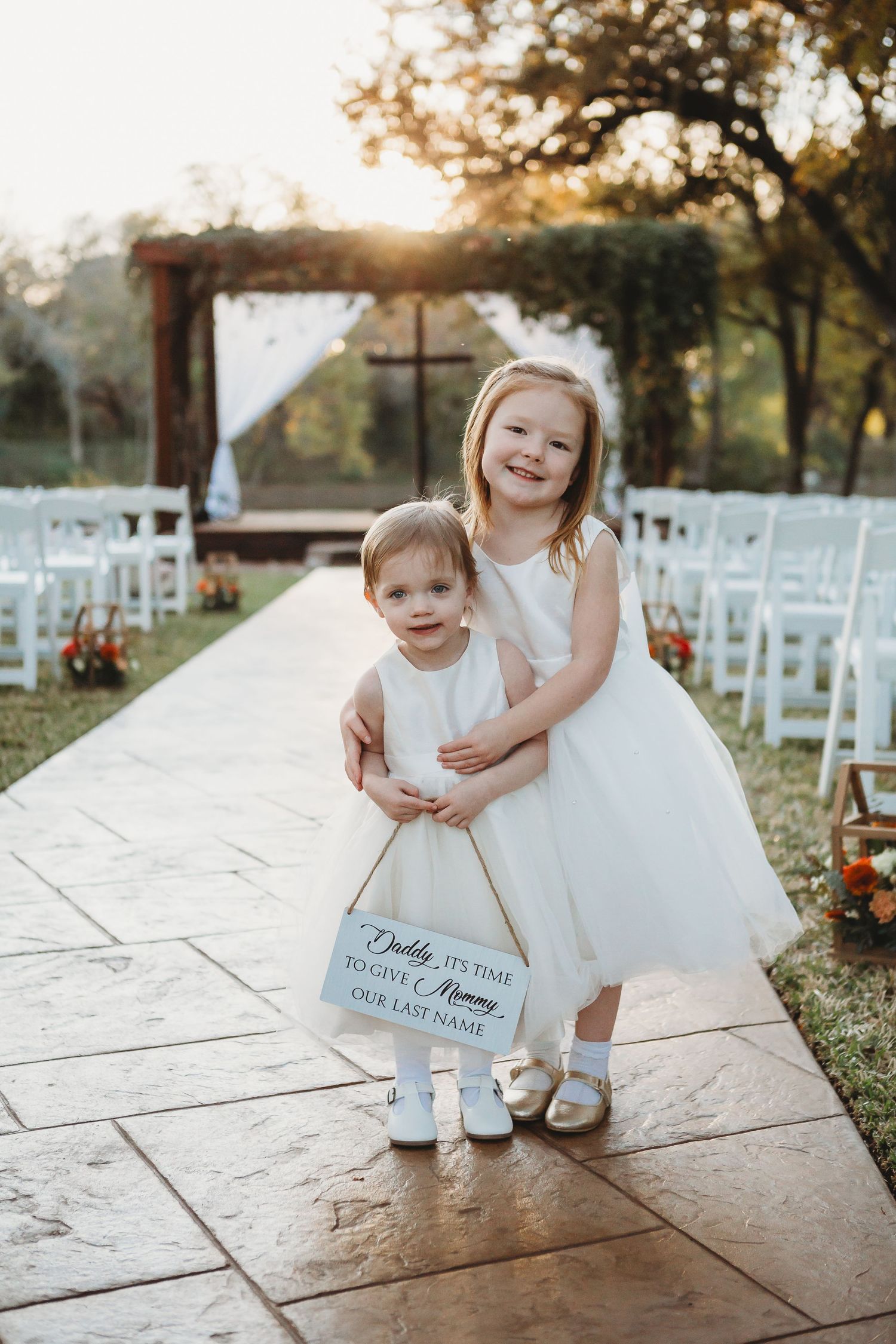 Two flower girls in white dresses stand together on stone path at outdoor wedding venue.