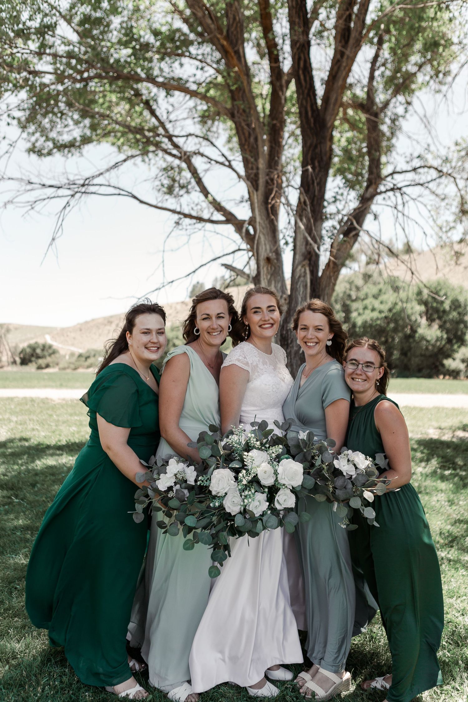Bridal party with white floral bouquets posing outdoors under a large tree.