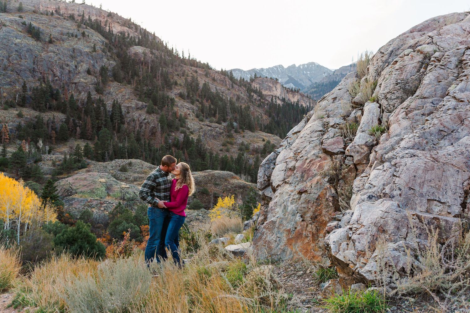Couple embracing near rocky cliff with scenic mountain valley view in background.