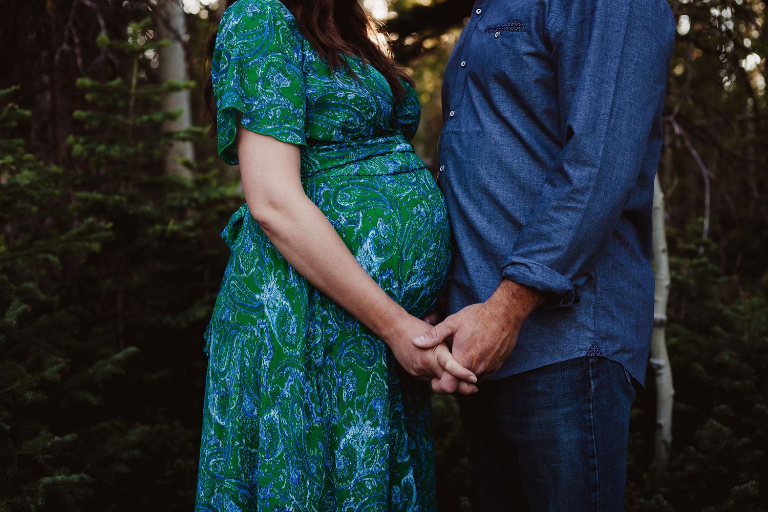 An intimate forest maternity photo showing a couple in navy and teal holding hands.