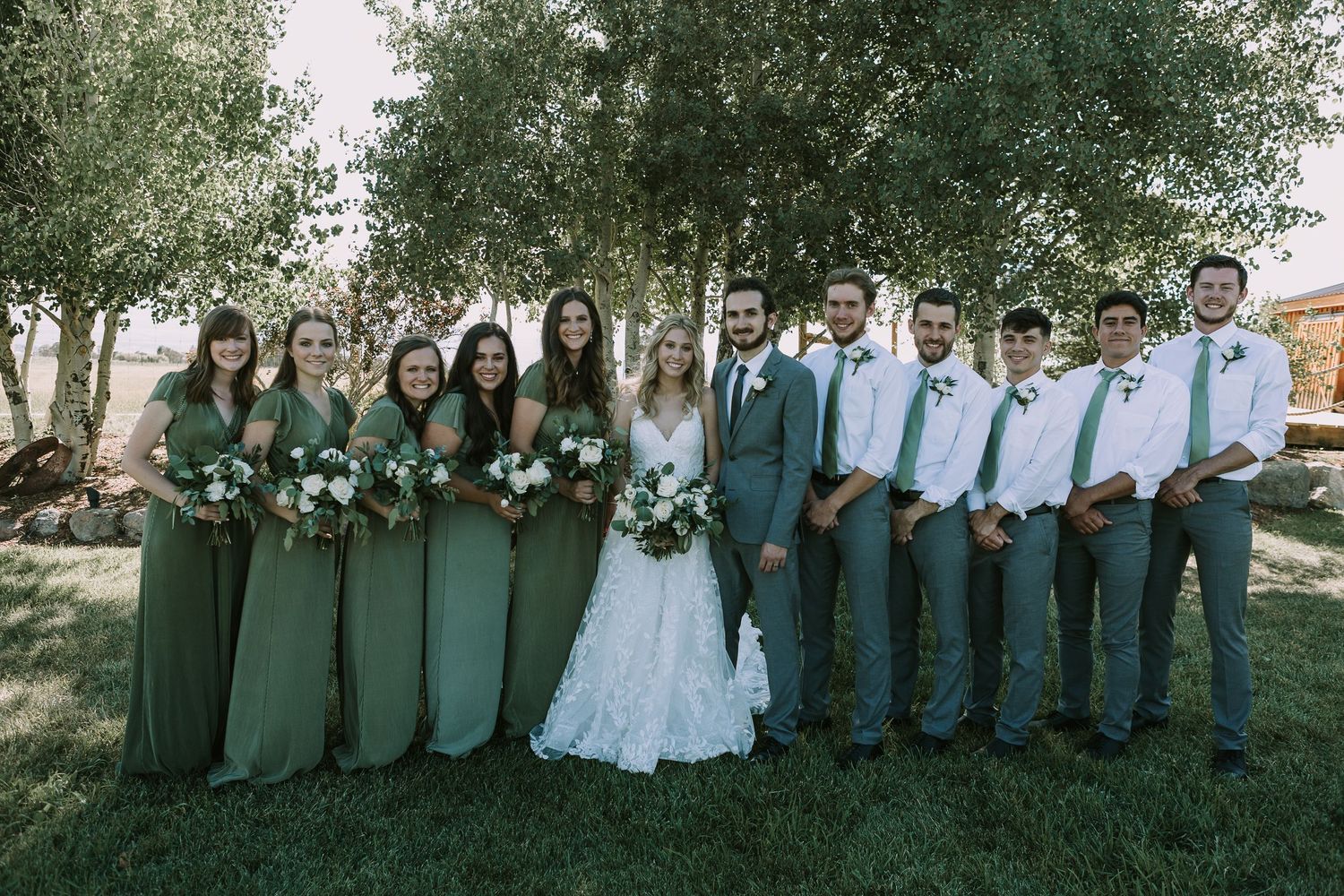 Wedding party poses outdoors with bridesmaids in emerald green dresses and groomsmen in gray suits.