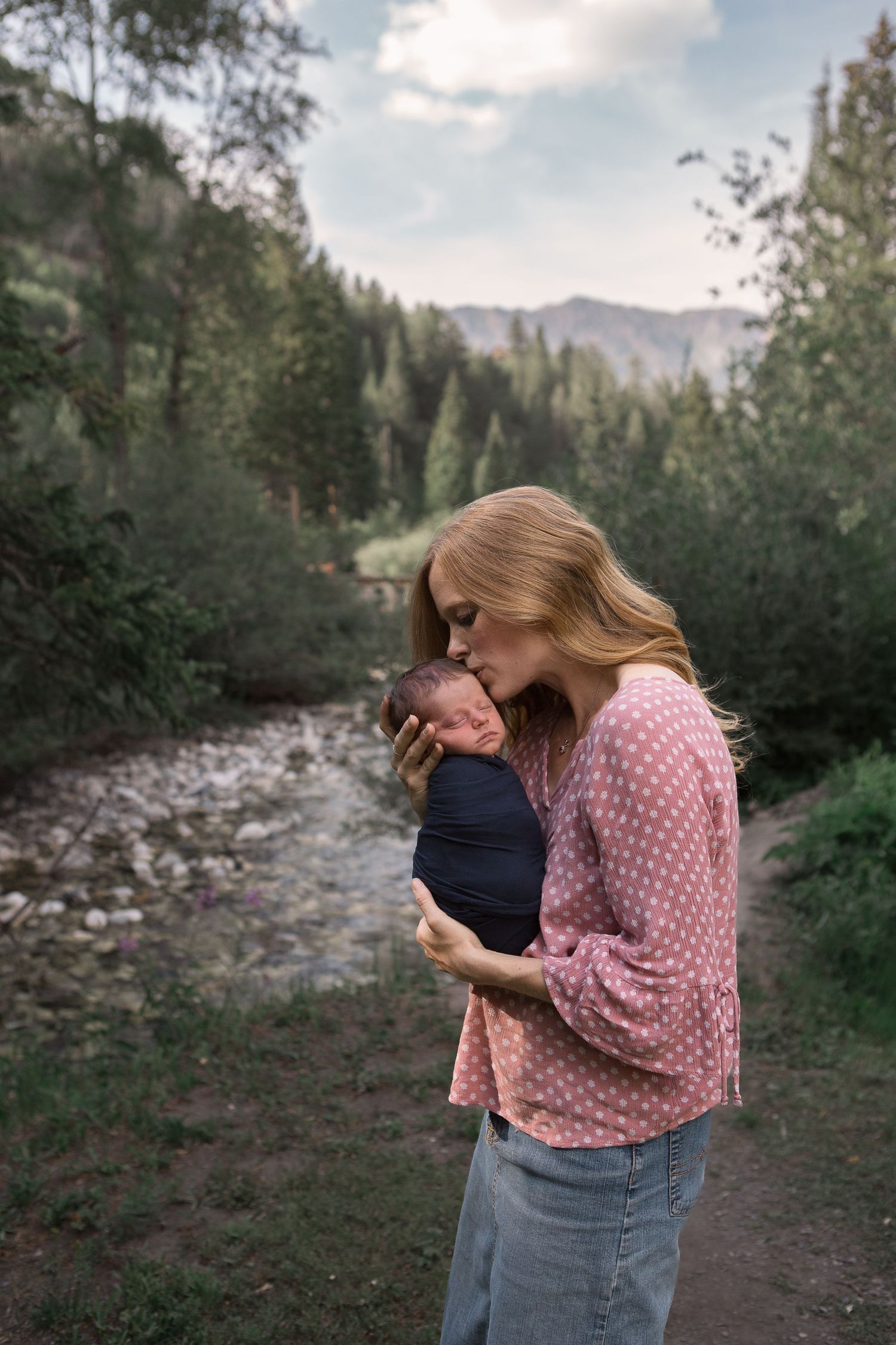 Gentle embrace with infant on mountain trail surrounded by pine trees and flowing creek.