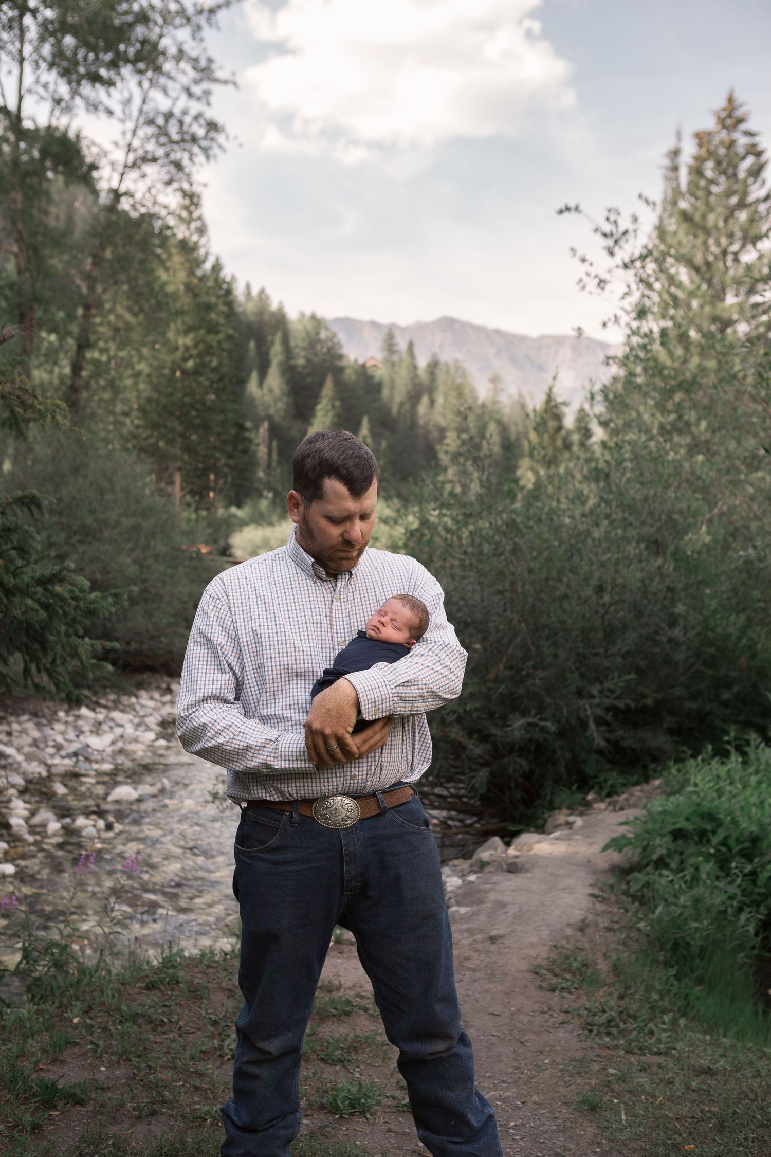 A person in a striped shirt holds a sleeping infant on a scenic mountain trail.